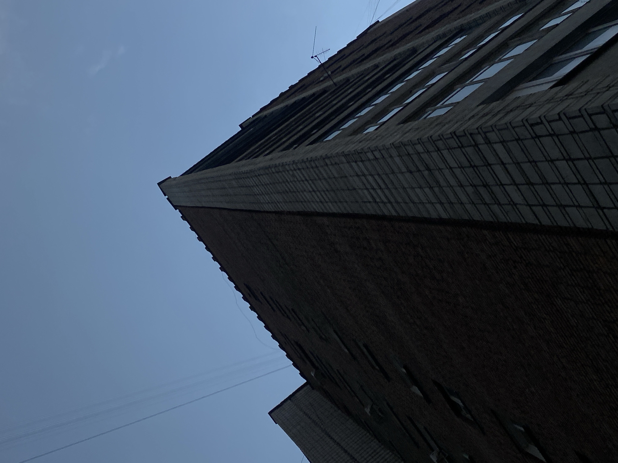 An extreme low-angle shot captures the corner of a multi-story brick building against a clear, pale blue sky. The building appears to be an apartment complex, with rows of windows visible on its facade. The brickwork on the right side of the building is a lighter grey, with a grid-like pattern, while the left side is constructed of darker, reddish-brown bricks. Several thin, dark lines, possibly electrical wires, stretch diagonally across the sky in the upper left quadrant of the image. A small, black television antenna is mounted on the side of the building near the top right. The image does not contain any people or evidence of activity. The perspective emphasizes the towering height of the building.
