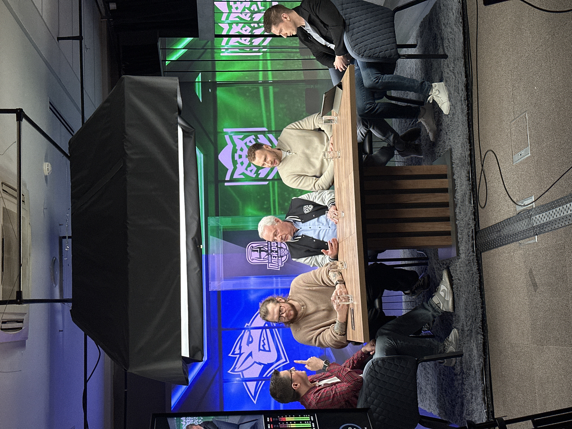 The image depicts a group of five men seated at a long, wooden table. They are positioned in front of a large screen displaying a logo in green and blue hues. The men are dressed in a variety of casual attire, including a red flannel shirt, a beige sweater, a varsity jacket, jeans, and sneakers. Two of the men have glasses. One man appears to be pointing at another. In front of them on the table are water glasses, a laptop and other items. Overhead, there is a black awning-like structure. A lighting fixture is visible, illuminating the scene. The environment seems to be a studio or broadcast setting. Location: Unknown, Unknown.