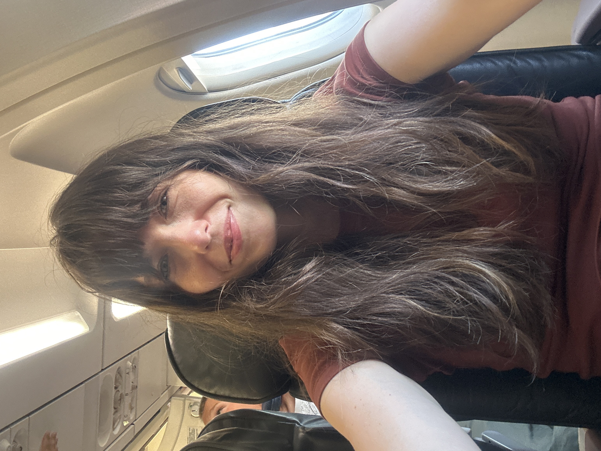 A woman with long, brown, wavy hair and bangs is seated on an airplane. She is wearing a maroon t-shirt and is taking a selfie, smiling at the camera. The background shows the interior of an airplane cabin, with seats, a window, and overhead compartments visible. Another person is partially visible in the seat behind her. The lighting suggests it is daytime. This image was taken inside Dalaman Airport, Turkey.