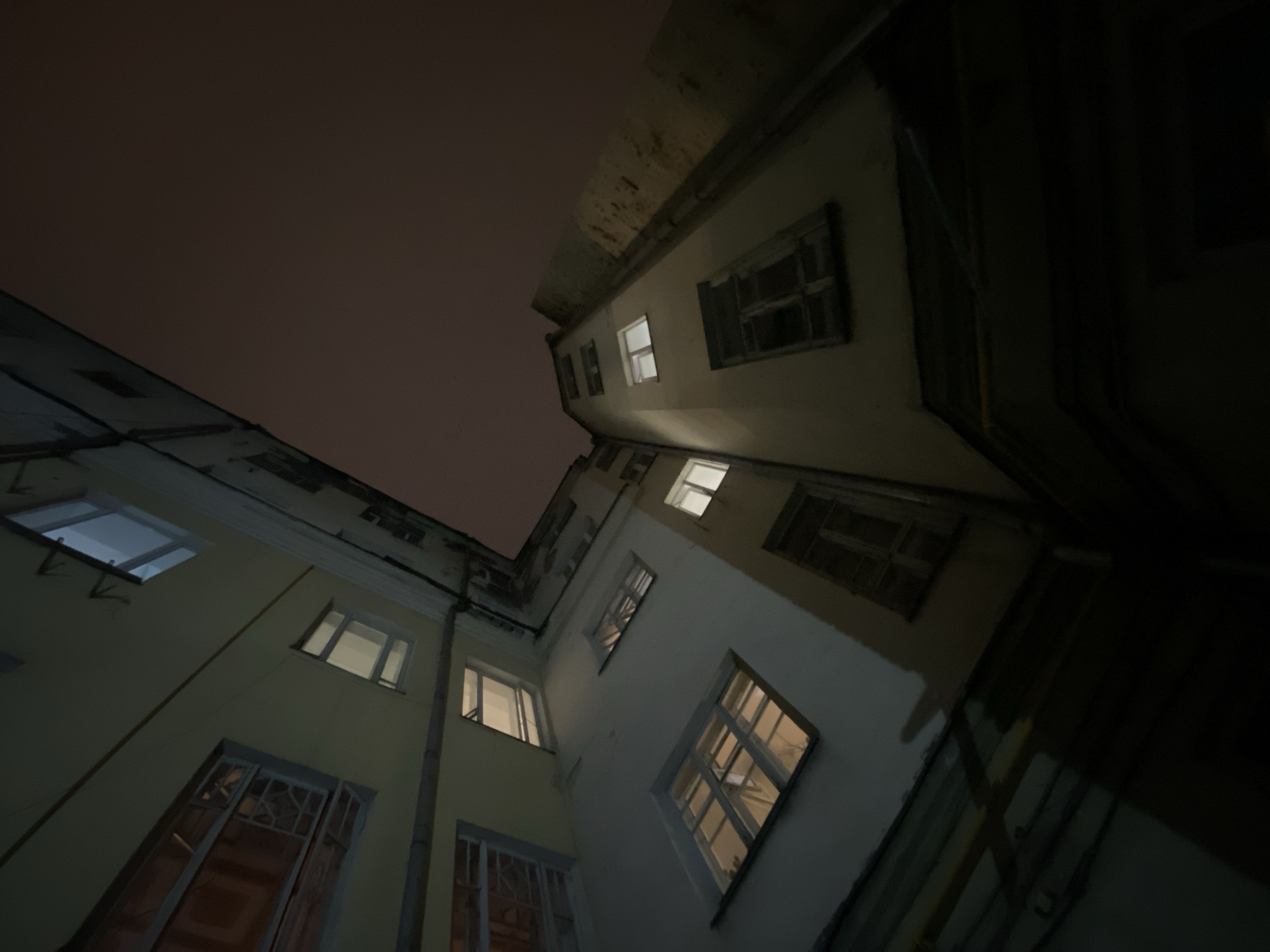 The image depicts a view looking up at the exterior walls of two buildings at night. The walls are painted in light shades, possibly pale yellow or beige, and have several windows of varying sizes. Some windows are lit from within, suggesting occupancy. The sky above is dark, indicating nighttime. The angle of the shot suggests the photo was taken from the ground, looking up into a courtyard or narrow space between the buildings. The architecture appears to be typical of older urban buildings, possibly located in Moscow, Russia. There are no people visible in the image.