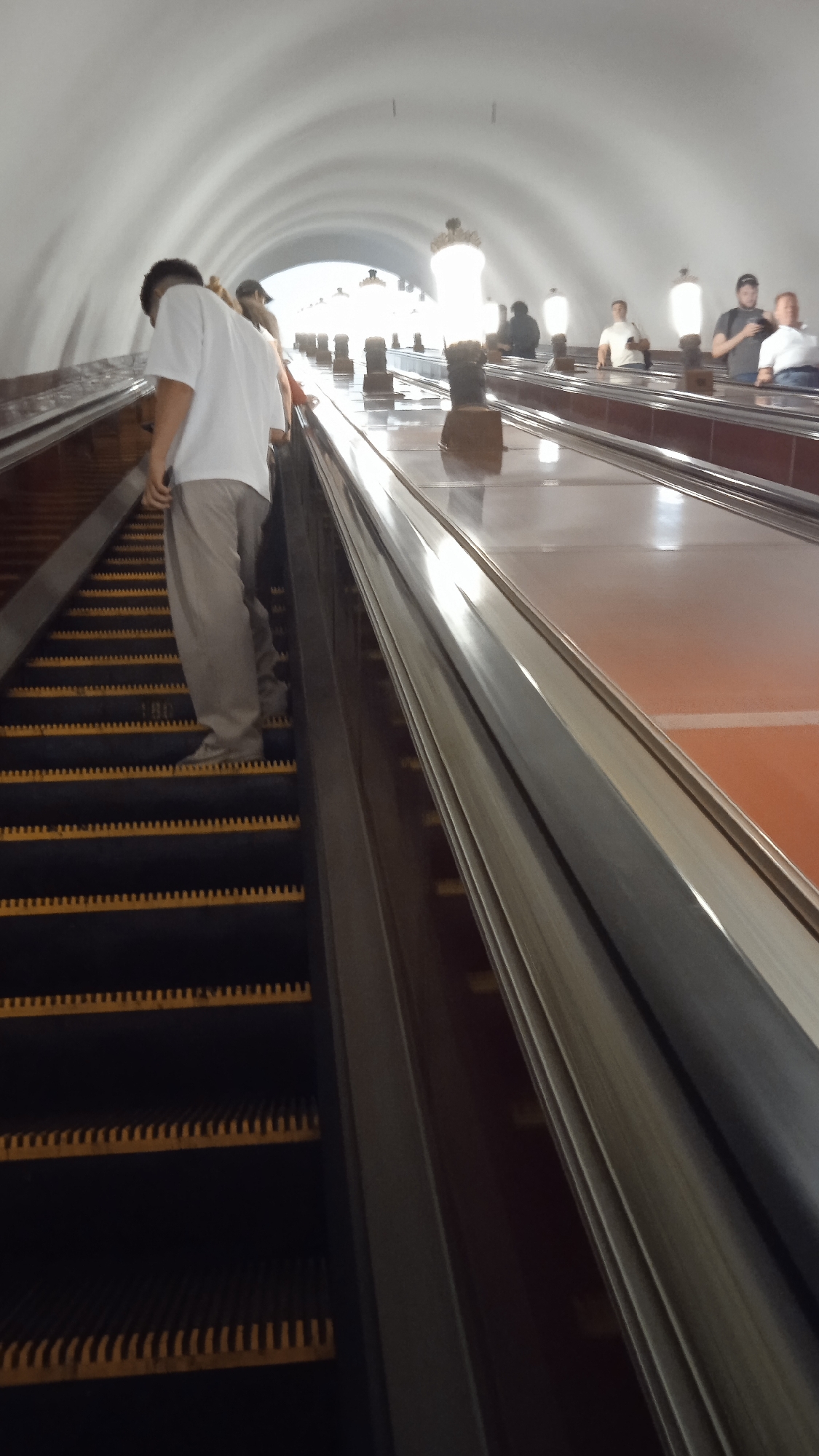 The image shows a broad, vaulted escalator tunnel with a group of people ascending. The escalator steps are black with yellow-lined edges. In the foreground, a person wearing a white t-shirt and light-colored pants is ascending on the left side of the escalator. Further up the escalator, several other individuals are visible, some standing and others ascending. The tunnel walls are white and curved, forming a wide arch overhead. The lighting appears to be from overhead fixtures and the bright light at the far end of the tunnel. The overall scene suggests a public transportation hub, likely a metro station due to the architectural style and escalator design. This architectural style is characteristic of Moscow's deep metro stations, many of which were built in the Soviet era and are known for their grandeur.