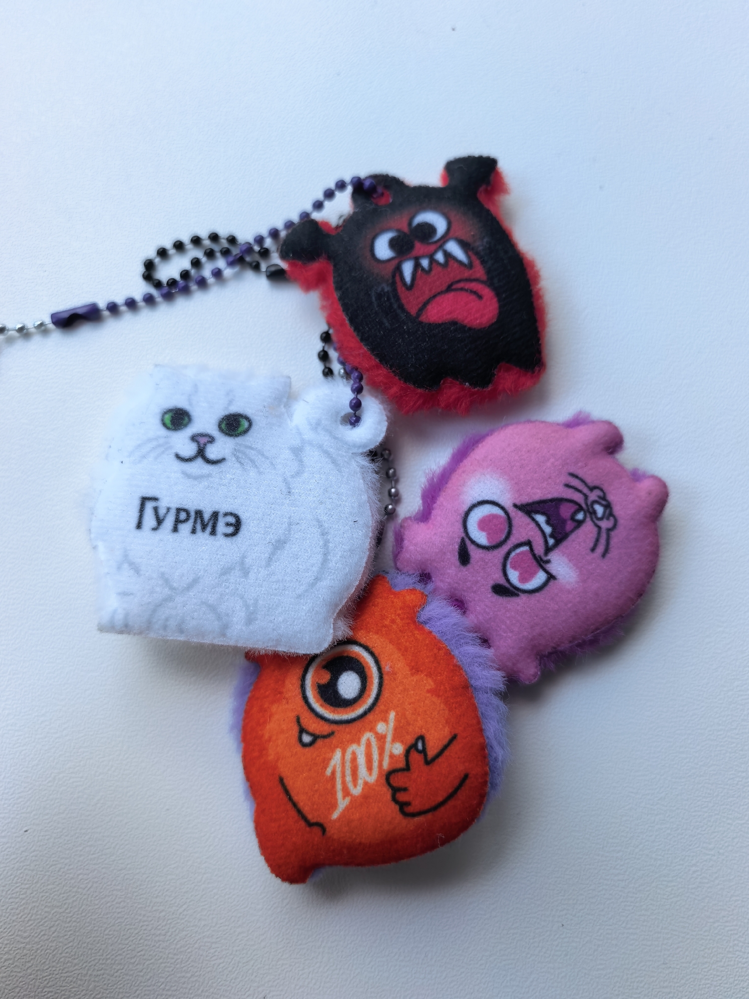 A close-up shot depicts four plush toy keychains arranged on a white surface. From left to right, there is a white plush cat with the Russian word "Гурмэ" printed on it; a black and red plush monster; a pink plush monster; and an orange plush monster with "100%" printed on it. The toys appear to be attached to each other with a ball chain, some of which are black. The photo was possibly taken in Sterlitamak, Russia, based on the name "Гурмэ" (Gourmet) being written in Russian.
