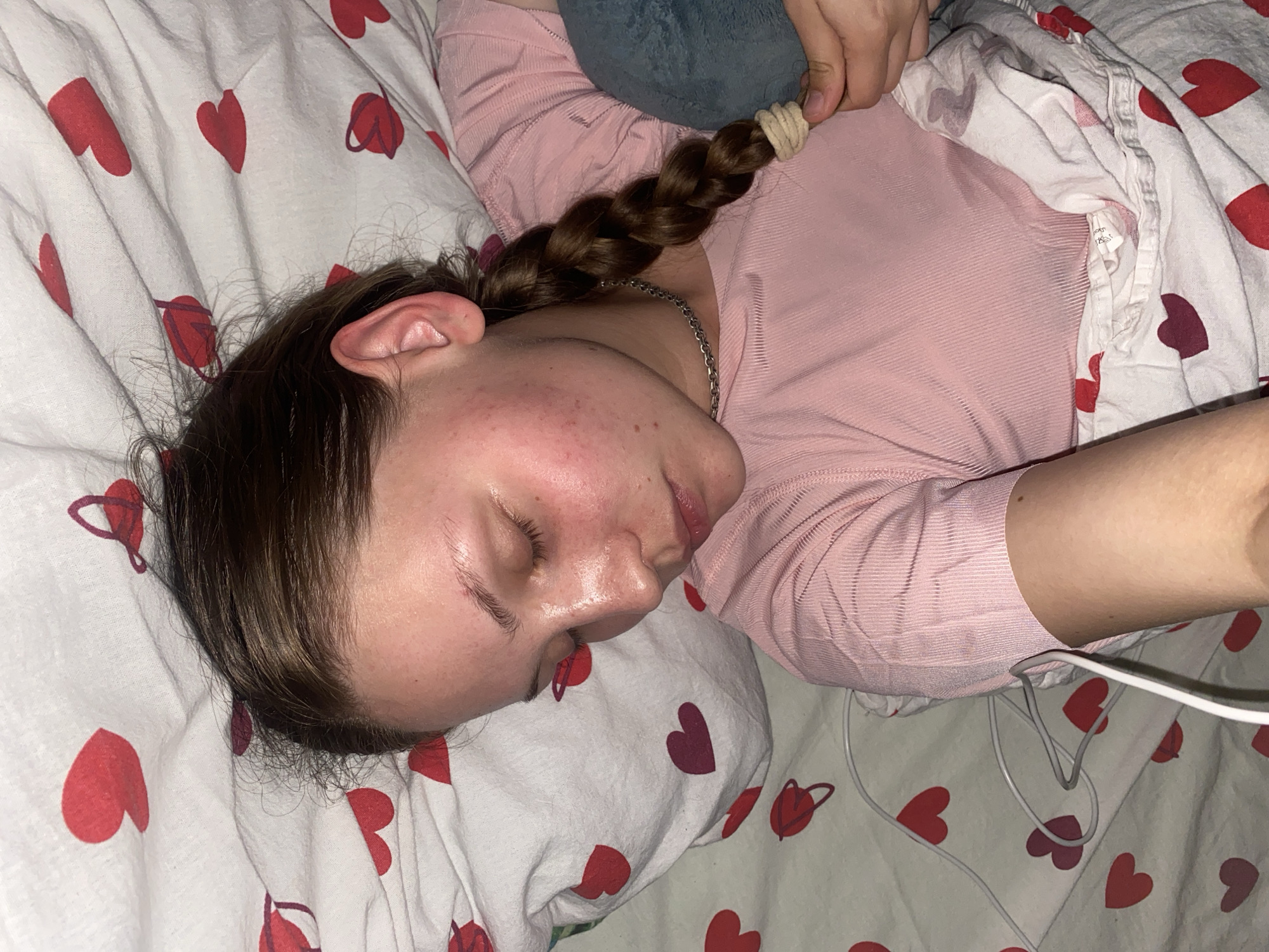 The media file shows a young person sleeping in a bed with a heart-patterned duvet. The person is lying on their side, facing left, with their eyes closed and their face resting on a pillow. They have dark brown hair braided into a single plait, which is secured with a beige elastic band. The person is wearing a pink, long-sleeved top. A greyish-blue plush toy is visible behind their head, partially covered by the duvet. White and grey wires are seen near the person's shoulder, trailing off the right side of the frame. The bedding has a repeating pattern of red and dark pink hearts on a white background. The lighting suggests it might be daytime or a well-lit indoor setting. There is no visible text in the image. The scene conveys a sense of rest and peaceful sleep. The location context provided is Вассята, Russia, which does not offer specific visual cues within the image itself.