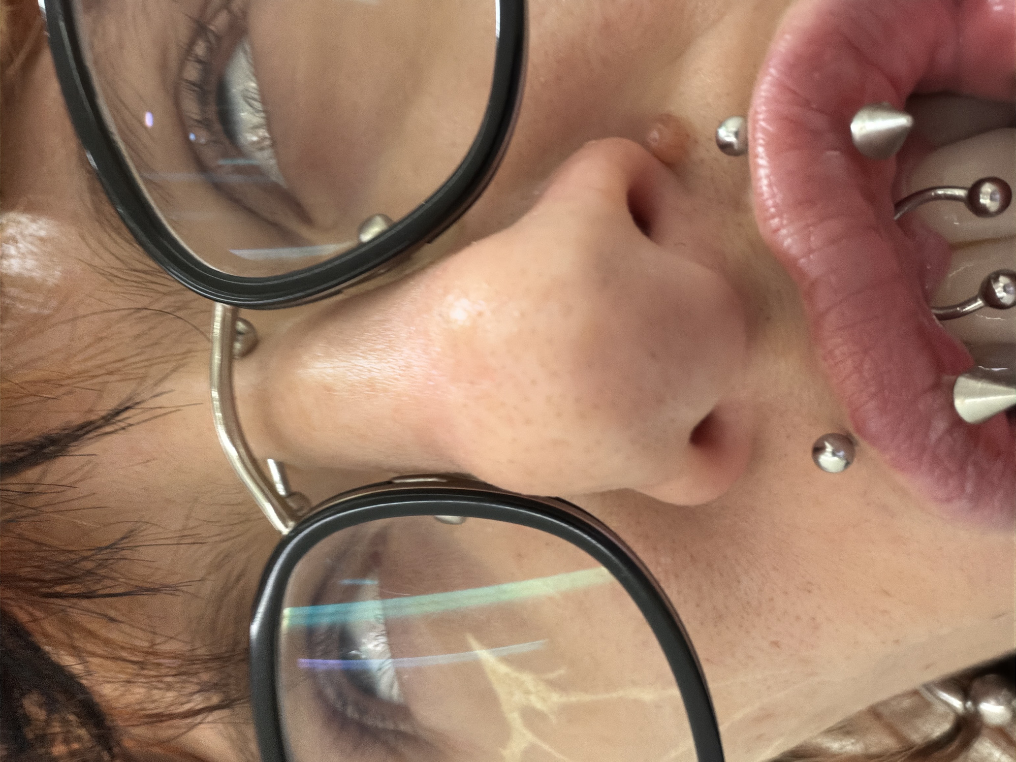 This close-up portrait presents a heavily cropped and rotated view of a person's face, oriented approximately 90 degrees counter-clockwise. The subject wears black-rimmed glasses, with distinct bright reflections across the lenses. Visible behind the lenses are parts of the eyes and eyelashes.
The most striking features are the numerous silver-toned piercings. A septum ring passes through the nose, accompanied by small stud piercings on either side of the nostrils. Around the mouth, several labret or lip piercings are visible, including one with a sharp, conical spike and others with spherical ends. The mouth is slightly open, revealing the tongue, which is adorned with multiple barbell-style piercings, and parts of the teeth.
The skin on the nose and surrounding areas shows visible pores and natural texture. A small, raised mole or bump is noticeable on the upper cheek near the left nostril (as perceived in an upright orientation). Wisps of dark, possibly light brown hair are visible along the left edge of the frame. The image offers an intimate, detailed examination of facial features and body modifications. No specific activity is depicted beyond posing for the photograph. The scene is likely an indoor setting in Busmantsi, Bulgaria. No text is visible.