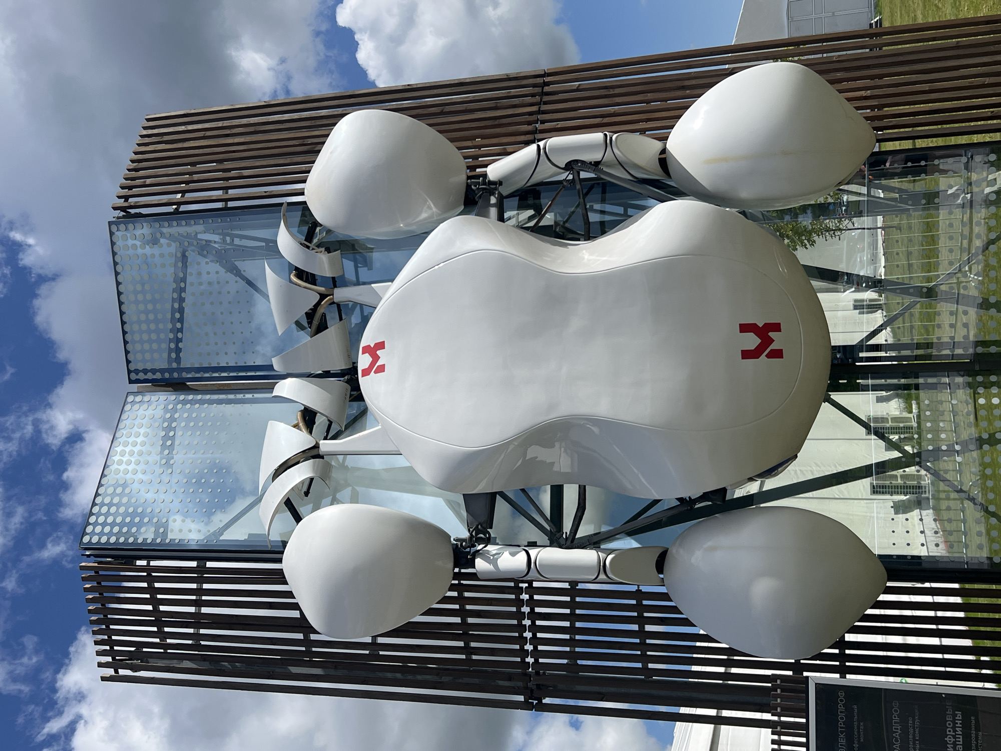 The image shows a large, white, bulbous structure with red logos resembling the letter "M" or a stylized "H" on its surface. This structure appears to be part of an outdoor art installation or sculpture. It is positioned in front of a modern building with a glass facade, which is framed by vertical wooden slats on either side. The sky above is blue with white clouds. In the foreground, to the left, is a sign with Cyrillic text, including the words "ЭЛЕКТРОПРОФ" and "РАСАДПРОФ". The overall scene suggests an exhibition or public space in Veliky Novgorod, Russia. No people are visible in the image.