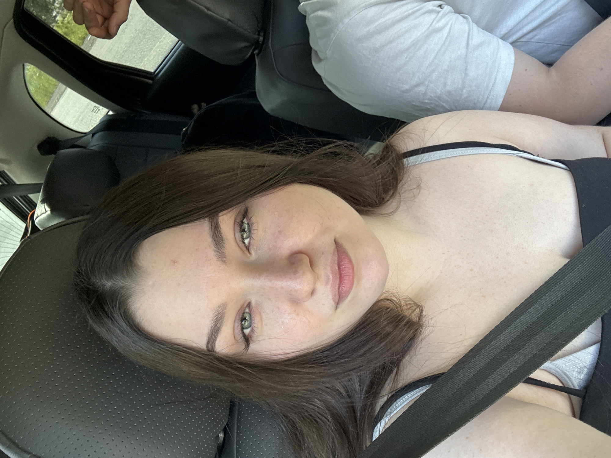 The image shows a young woman with long brown hair and green eyes, wearing a black tank top and a grey bra, with a seatbelt across her chest. She is seated in the front passenger seat of a car, looking directly at the camera. The interior of the car is visible, including the headrest of the driver's seat and a portion of the passenger seat. The window on the right shows a blurry view of trees and a road, suggesting the car is in motion. Another person's arm, wearing a light-colored t-shirt, is visible on the right side of the frame. The scene is lit by natural daylight. There is no indication of the city of Pesterevo, Russia.