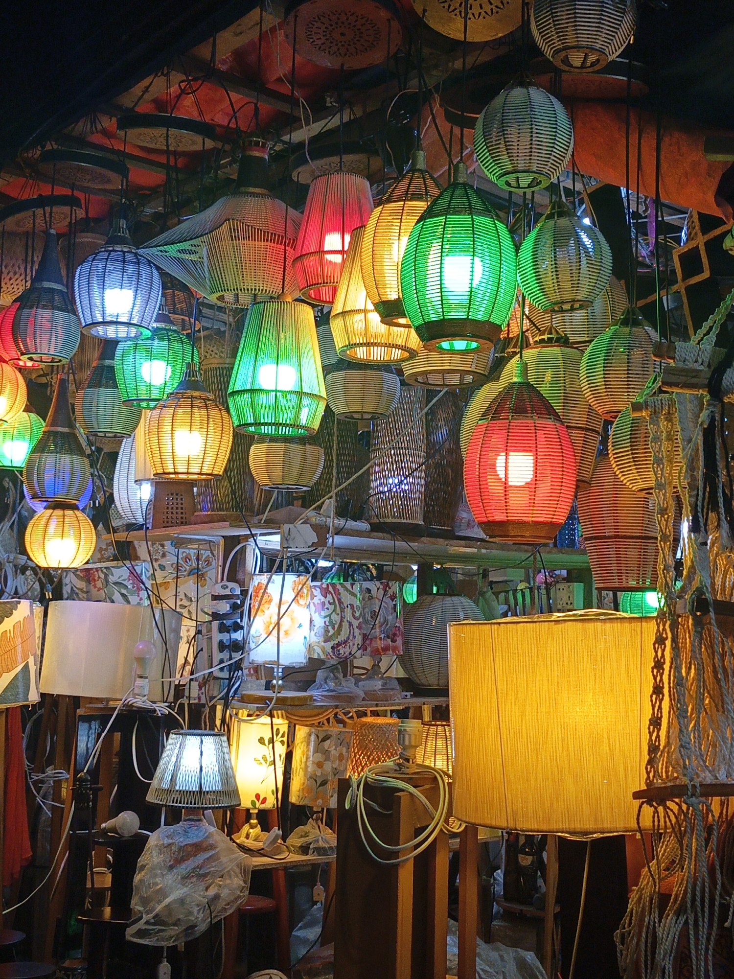This vibrant image captures a bustling display of numerous lamps and light fixtures, likely within a market stall or shop in Dhaka, Bangladesh. The scene is dominated by a dense arrangement of hanging lamps, crafted primarily from woven natural materials like rattan or bamboo. These shades come in various shapes—globular, conical, and cylindrical—and emit a spectrum of brilliant colors, including red, green, blue, yellow, and white, creating a kaleidoscope effect.
Below the suspended lights, the lower section of the image reveals a more cluttered environment with an array of table lamps and possibly a floor lamp. Some of these feature fabric shades, occasionally adorned with floral or geometric patterns. Wires crisscross the space, connecting various bulbs and fixtures. The ground level displays wooden stands, additional lamp bases, and miscellaneous items, some covered in plastic.
No people are visible, suggesting the focus is entirely on the merchandise. The artificial illumination from the lamps is the primary light source, indicating either an indoor setting with limited natural light or a nighttime scene, characteristic of a lively evening market. The handcrafted appearance of many lamps, coupled with the overall dense display, hints at a local artisan market or a specialized lighting store. The atmosphere is energetic and warm, filled with the glow of myriad lights. No discernible text is visible within the frame.