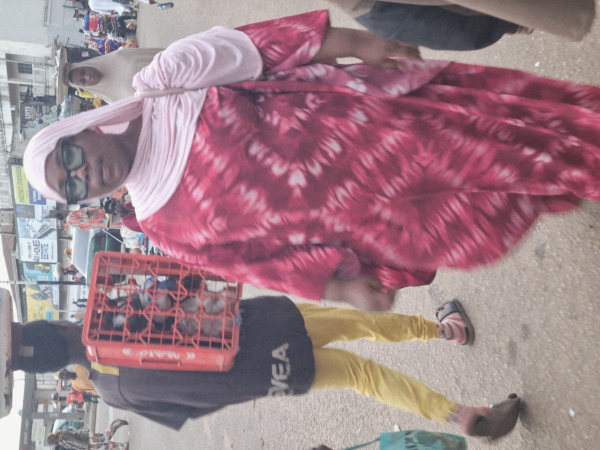 This is a vertically oriented, medium shot of people in an outdoor marketplace in Jos, Nigeria. The image is slightly blurry and appears to be taken at eye level. In the foreground, a person wearing a vibrant pink and white tie-dye dress and a light pink hijab is the most prominent subject. They are wearing glasses and looking slightly to the left. Below them, another person is wearing a dark gray shirt with the word "VEA" visible in white lettering, and bright yellow pants. Their feet are visible, one wearing a sandal with pink and white stripes. A red plastic crate filled with what appear to be bottles is held in front of them. In the background, more people are visible, some with head coverings, and market stalls with goods are suggested. There are also glimpses of buildings and signs. The ground appears to be a paved surface. The overall impression is a bustling market scene with a focus on the individuals in the foreground. The lighting suggests it is daytime.