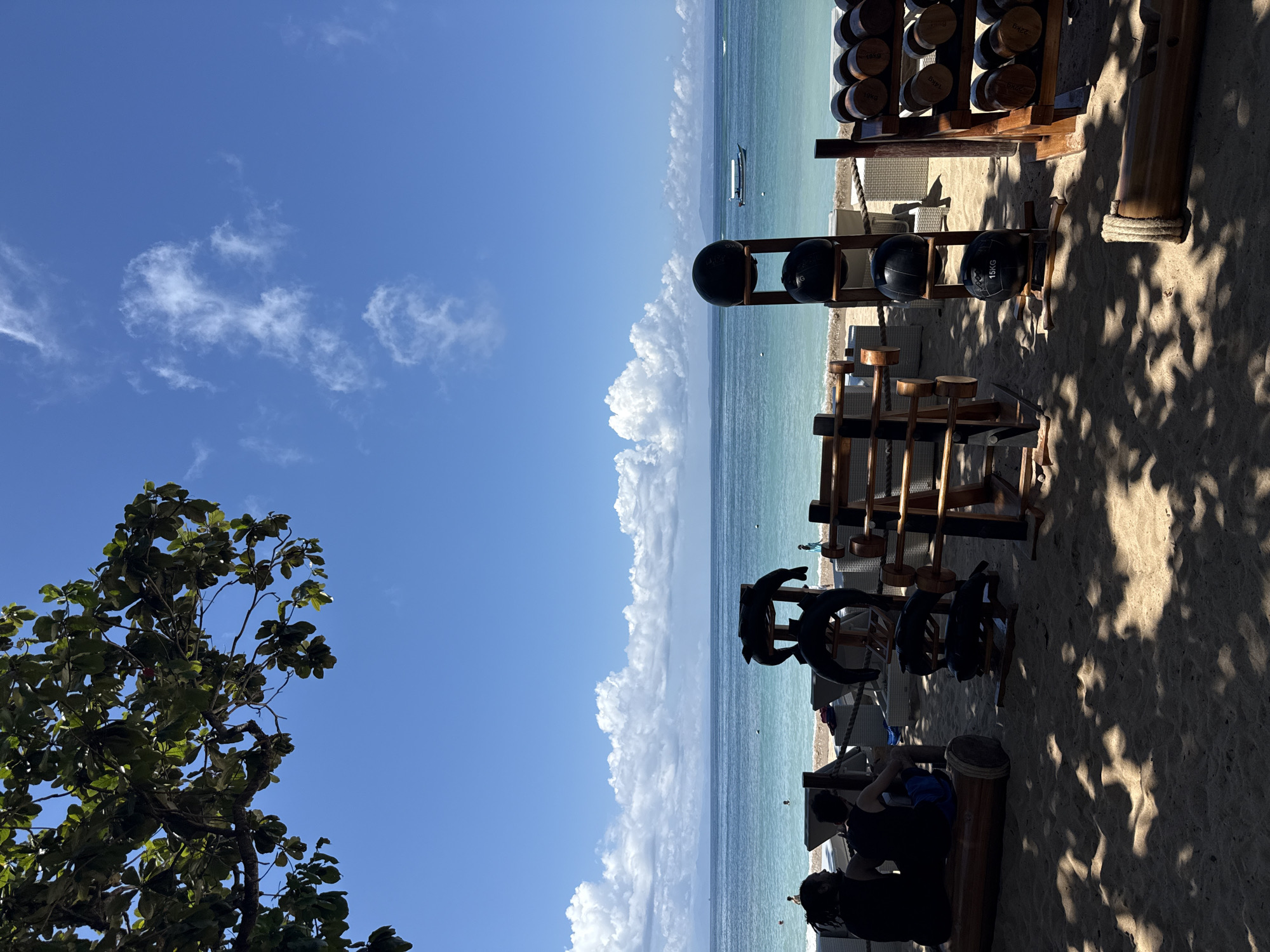 The image captures an outdoor gym setting on a beach in Nusa Dua, Indonesia. Various exercise equipment, including wooden dumbbells, medicine balls, and wooden mallets, are arranged on racks. A tree with green leaves is present in the lower left corner, and the beach stretches towards the turquoise ocean under a sky with scattered clouds. In the foreground, individuals are present, but obscured by shadows and the orientation of the photograph.