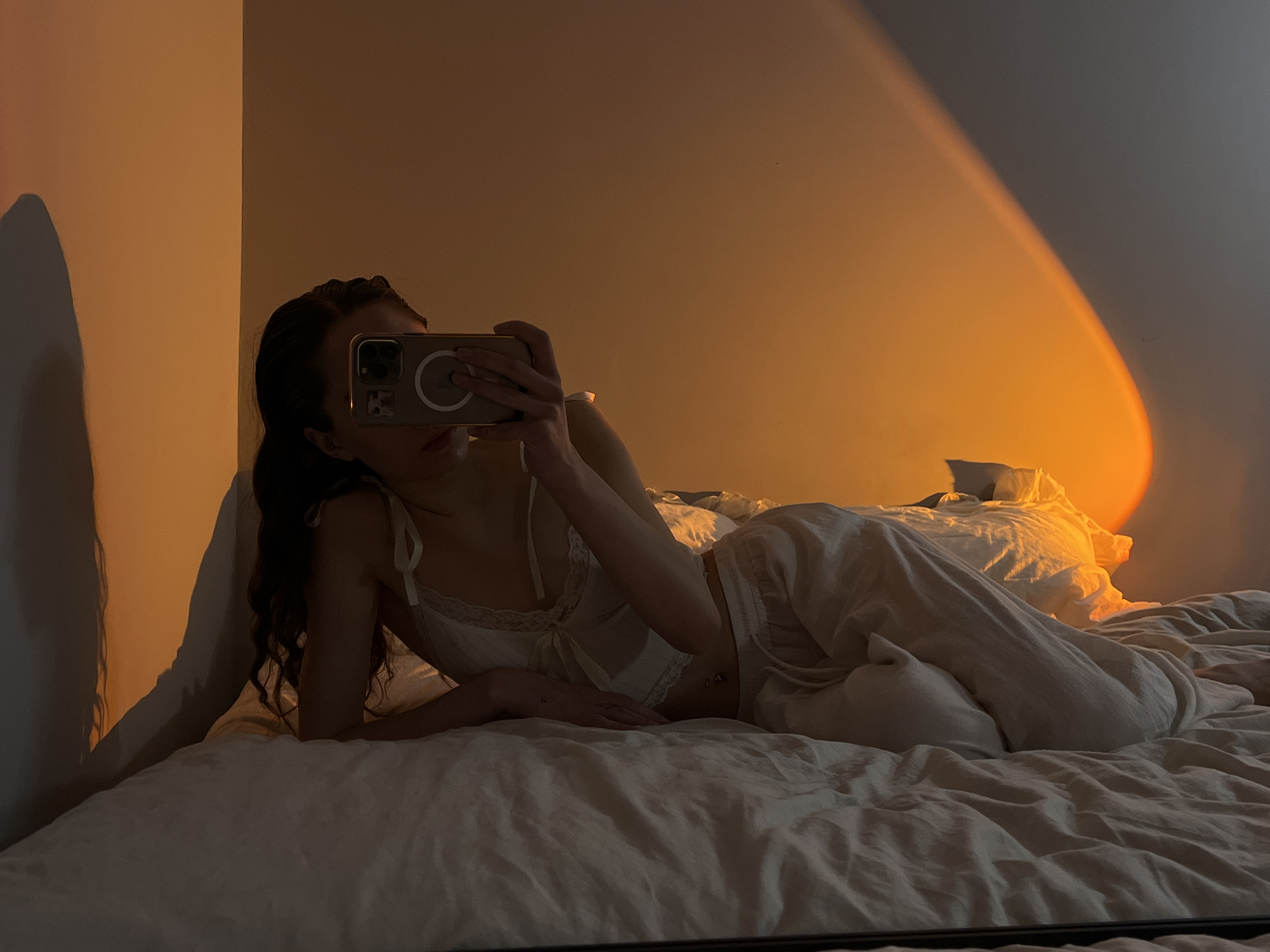 A young woman is lying on a bed, taking a selfie with her smartphone. She is wearing a white camisole and loose pants. The lighting is warm, with a golden hue illuminating the wall behind her. The bed is covered in crumpled sheets. The location is not evident from the image. The city of Ternopil, Ukraine, cannot be determined from this image.