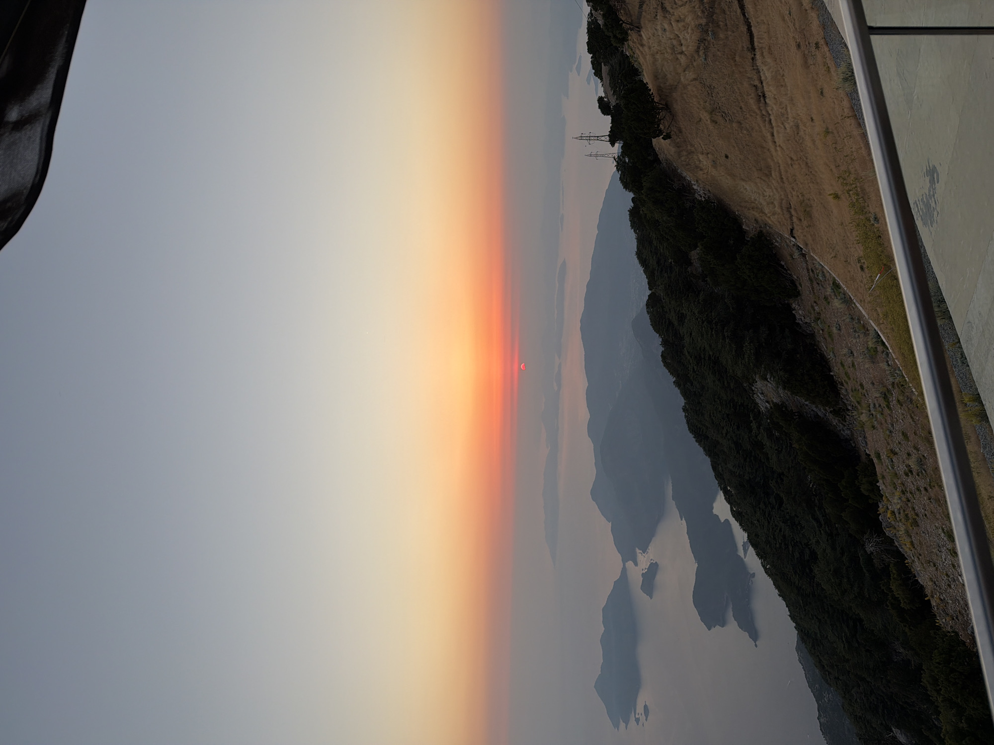 The image shows a panoramic view of Fethiye, Turkey, at sunset. The sun is a bright orange orb low on the horizon, casting a warm glow over the sky and water. Islands and mountains are visible in the distance, silhouetted against the colorful sky. The foreground features a steep, tree-covered hillside and a paved area with a railing, suggesting a viewpoint or a balcony overlooking the landscape.