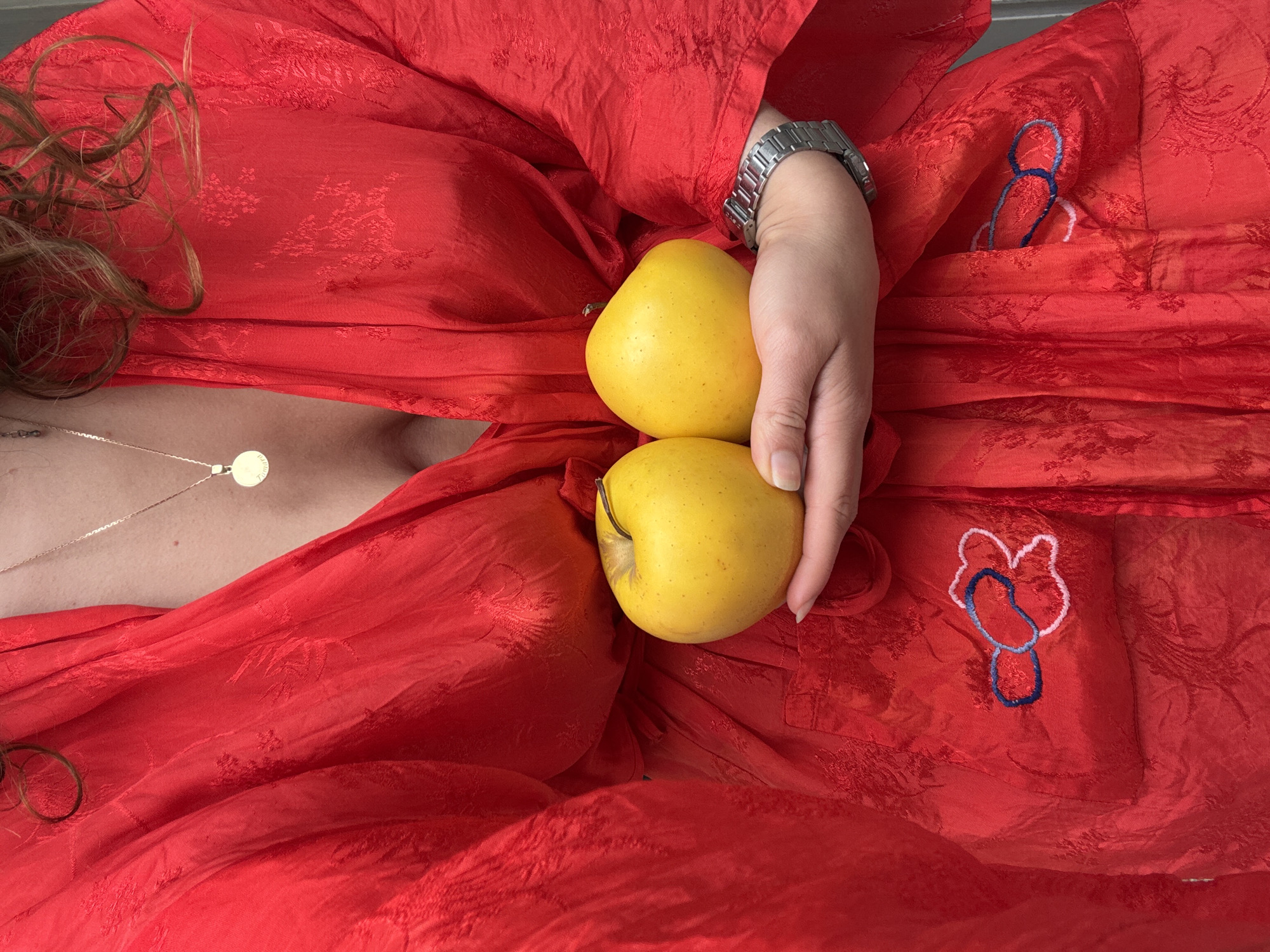 The image shows a close-up of a person's torso, wearing a vibrant red robe with a subtle pattern. The robe is open, revealing a glimpse of a necklace with a circular pendant. The person is holding two yellow apples in their hands, which are positioned in front of the robe. The robe has an embroidered detail on the chest area, featuring blue and pink floral elements. A silver watch is visible on the person's left wrist. The background is out of focus and suggests an indoor setting. There is no indication of the specific location within Tbilisi, Georgia, or any other contextual environmental details.