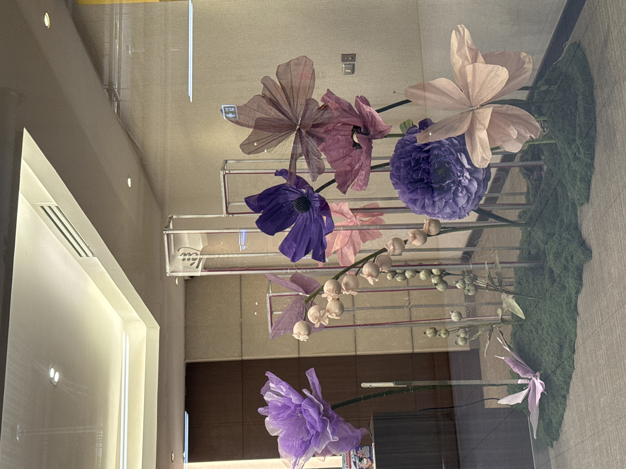 This media file depicts an artistic floral arrangement, likely an indoor display, as suggested by the reflective surfaces and architectural elements. The primary subjects are artificial flowers in various shades of purple, pink, and beige, arranged in a cascading manner. These flowers have a delicate, translucent appearance, resembling paper or silk. Some are large and resemble anemones or peonies, while others are smaller and bell-shaped, resembling lily of the valley. The arrangement is set against a backdrop of what appears to be textured wallpaper and a reflective surface, possibly a glass partition or window, which also reflects the surrounding environment, including lights and interior architecture.
The arrangement is supported by a metallic structure, featuring horizontal bars that create a tiered effect. One portion of the display includes a bed of green moss, adding a naturalistic element. The lighting appears to be artificial, casting soft illumination and creating subtle shadows. There are no people visible in the frame. The overall mood is serene and artistic, emphasizing the beauty and craftsmanship of the floral display.
The location context provided places this scene in Nong Prue Subdistrict, Thailand. However, no explicit visual cues within the image definitively confirm this location, as the interior setting is generic. The time of day is not discernible. No weather conditions are visible. There is some text visible in the bottom right corner of the original image, on what appears to be a small placard or advertisement, but it is too blurry to read.