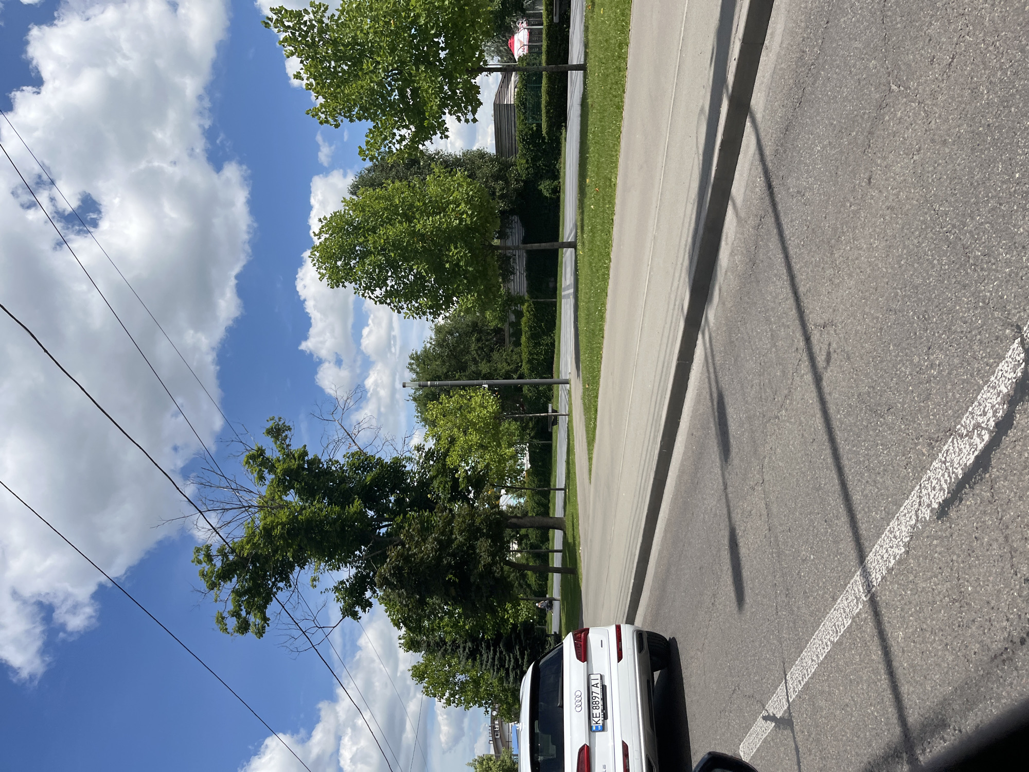 The image depicts an outdoor scene on a sunny day, likely in the late morning or early afternoon given the bright sunlight and strong shadows. The camera is positioned at an angle, looking down and to the side, as if from a vehicle.
The main subjects are a white Audi SUV, partially visible from the rear, and a row of trees lining a paved pathway or sidewalk. The car has a license plate with visible text: "KE 8897 AT". Beyond the pathway is a grassy area with more trees and some buildings in the background. The sky is a clear blue with scattered white cumulus clouds. Power lines are also visible crisscrossing the sky, suggesting an urban or suburban setting.
The scene appears to be a quiet street or park area. There are no prominent human figures visible in the immediate foreground or mid-ground, although a very small figure can be seen in the distance near the trees. The overall atmosphere is one of a pleasant, ordinary day. The location context provided is Dnipro, Ukraine. The weather is clear and sunny.