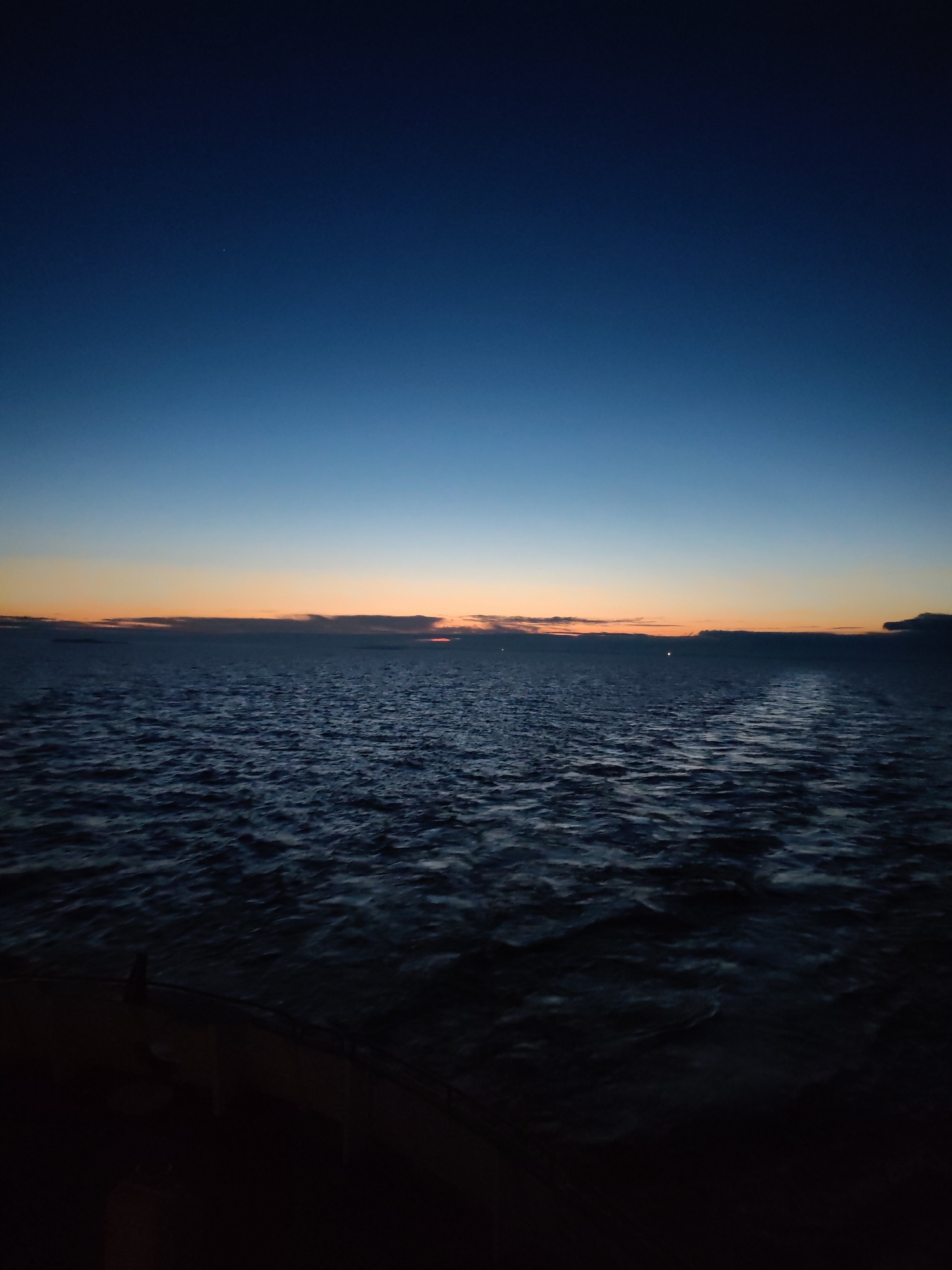 The image depicts a body of water, possibly a sea or a large lake, at dusk or dawn. The sky transitions from a deep blue at the top to orange near the horizon. There are some clouds on the horizon. The water is dark with visible ripples. The foreground is obscured and dark. The location is Unknown, Russia. There are no people visible.