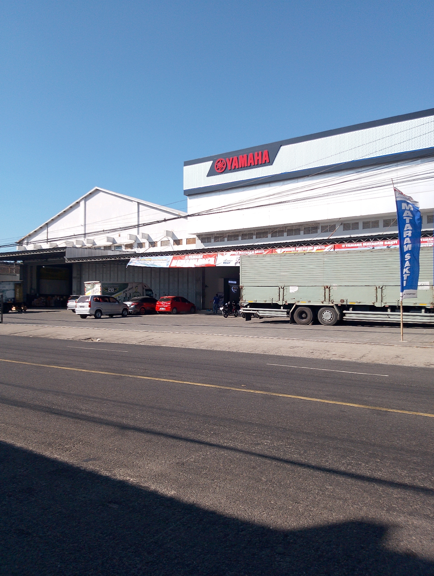 The image depicts a street scene in Guyangan, Indonesia, dominated by a Yamaha dealership. A large, white building with the Yamaha logo prominently displayed across its facade serves as the central subject. Adjacent to the dealership, a warehouse-like structure with a large opening is visible. In front of the dealership, several cars, including a silver hatchback, a red sedan, and a red smaller car, are parked. A light green truck with a covered bed is positioned on the road in front of the dealership, alongside a motorcycle. A blue flag with white lettering, partially obscured, stands vertically on a pole. The street in the foreground is asphalt with yellow and white markings, and shadows are cast across it. The sky is clear blue.