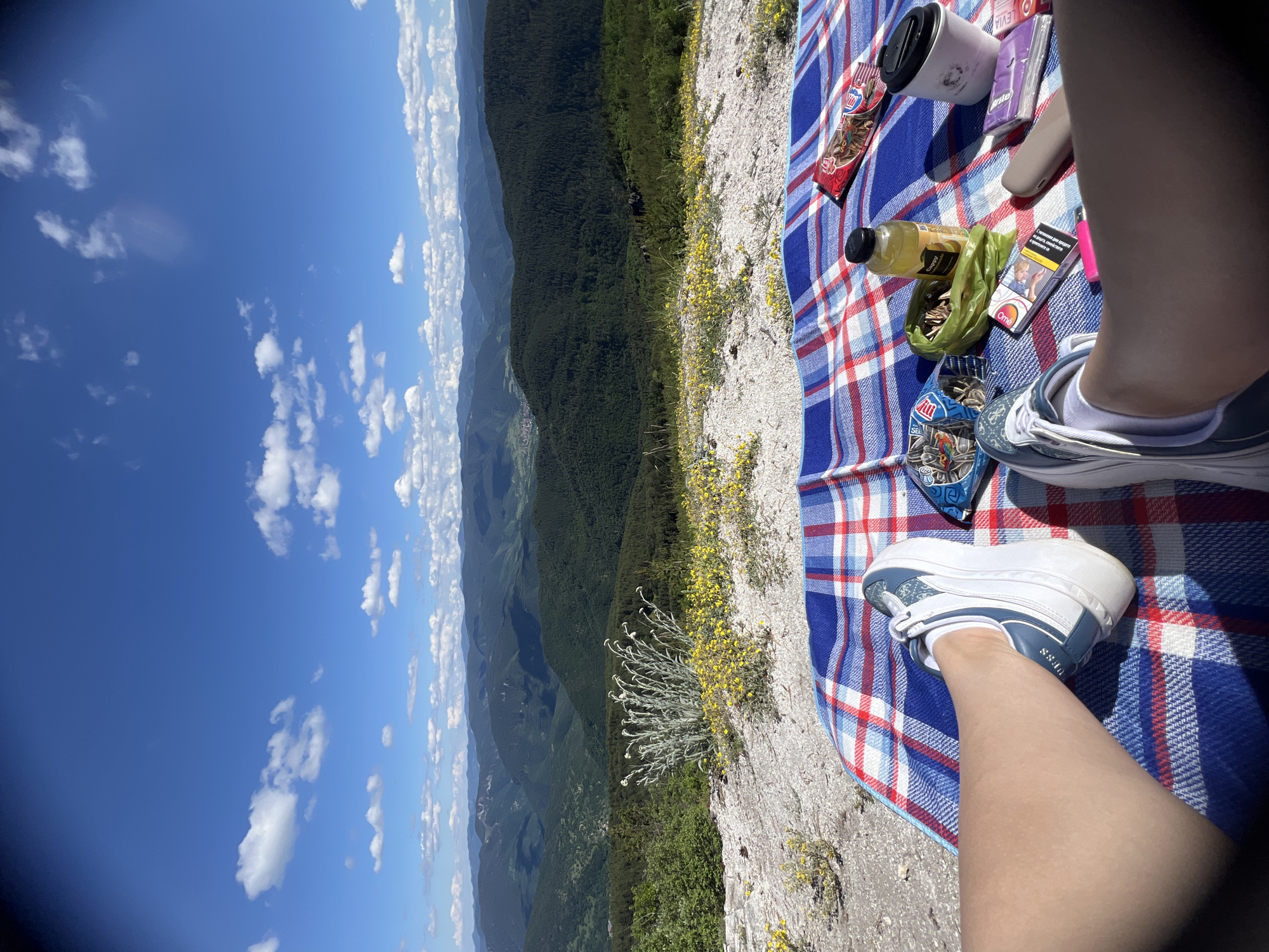 The image shows a first-person perspective of someone sitting on a red, white, and blue checkered blanket at a scenic overlook. The view is of rolling, green, forested mountains under a bright blue sky with scattered white clouds. In the foreground, on the blanket, are various items including a bottle of yellow liquid, a pack of sunflower seeds, a coffee cup, a pack of cigarettes, a pink lighter, and a beige object that appears to be a vape pen. The person is wearing white sneakers with blue accents and white socks. The ground beside the blanket is rocky with sparse yellow flowers and a bush with light-colored, feathery foliage. The location appears to be a natural, elevated area, possibly a mountain or hilltop, offering a panoramic view of a valley and further mountain ranges. There is no indication of it being in the city of Kosovo, Bulgaria.