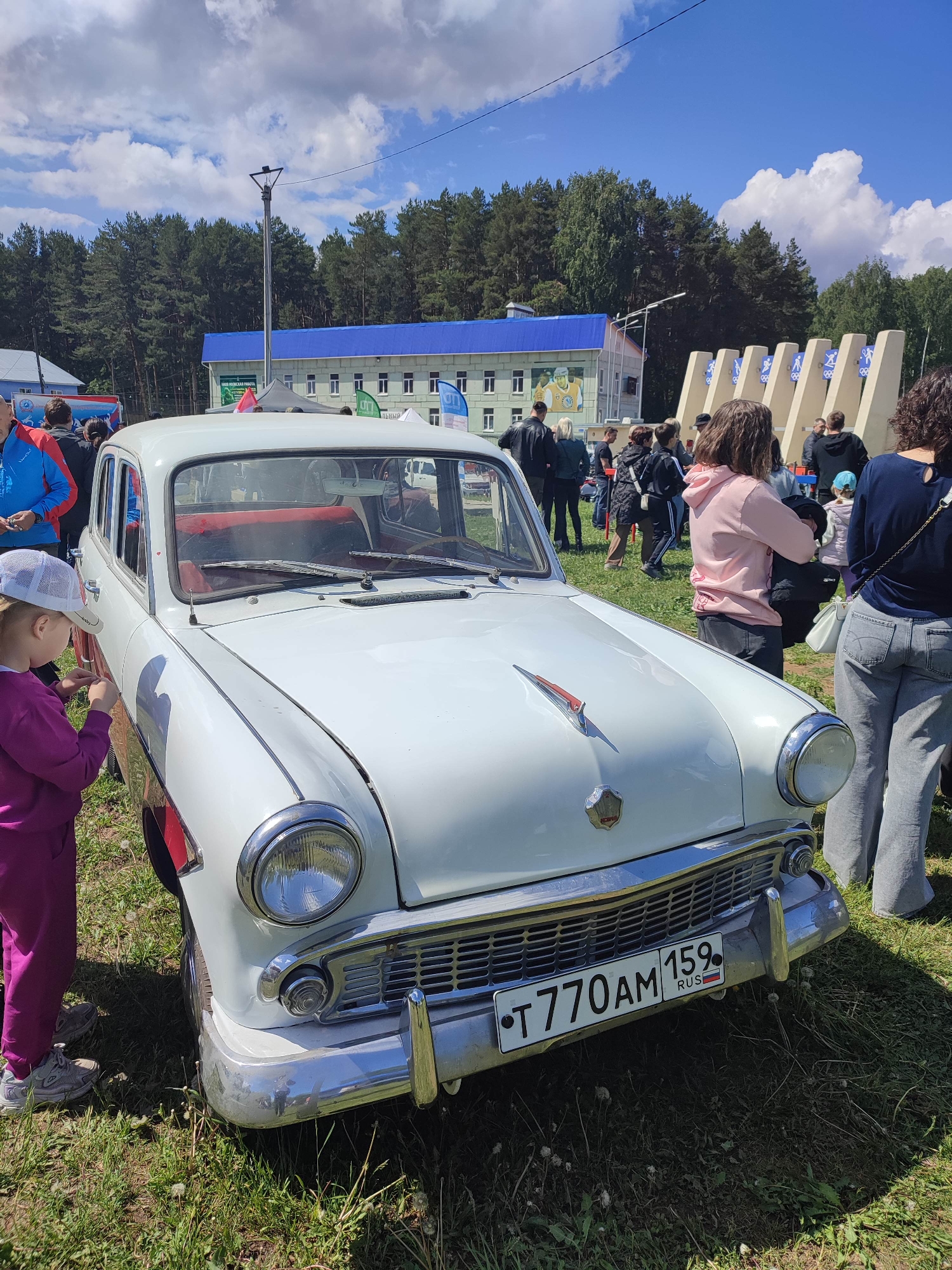 The image shows a white vintage car, presumably a Moskvich, parked on a grassy area with people gathered around. The car has a red and silver ornament on the hood and a Russian license plate reading "T770AM 159 RUS". To the left of the car stands a young girl in a pink jumpsuit and a white cap. To the right, a woman in jeans and a blue shirt stands facing away from the camera. In the background, there are more people, a building with a blue roof and white windows, a row of monument-like structures, and trees under a sunny, slightly cloudy sky. The location appears to be an outdoor event or car show in an unspecified city.