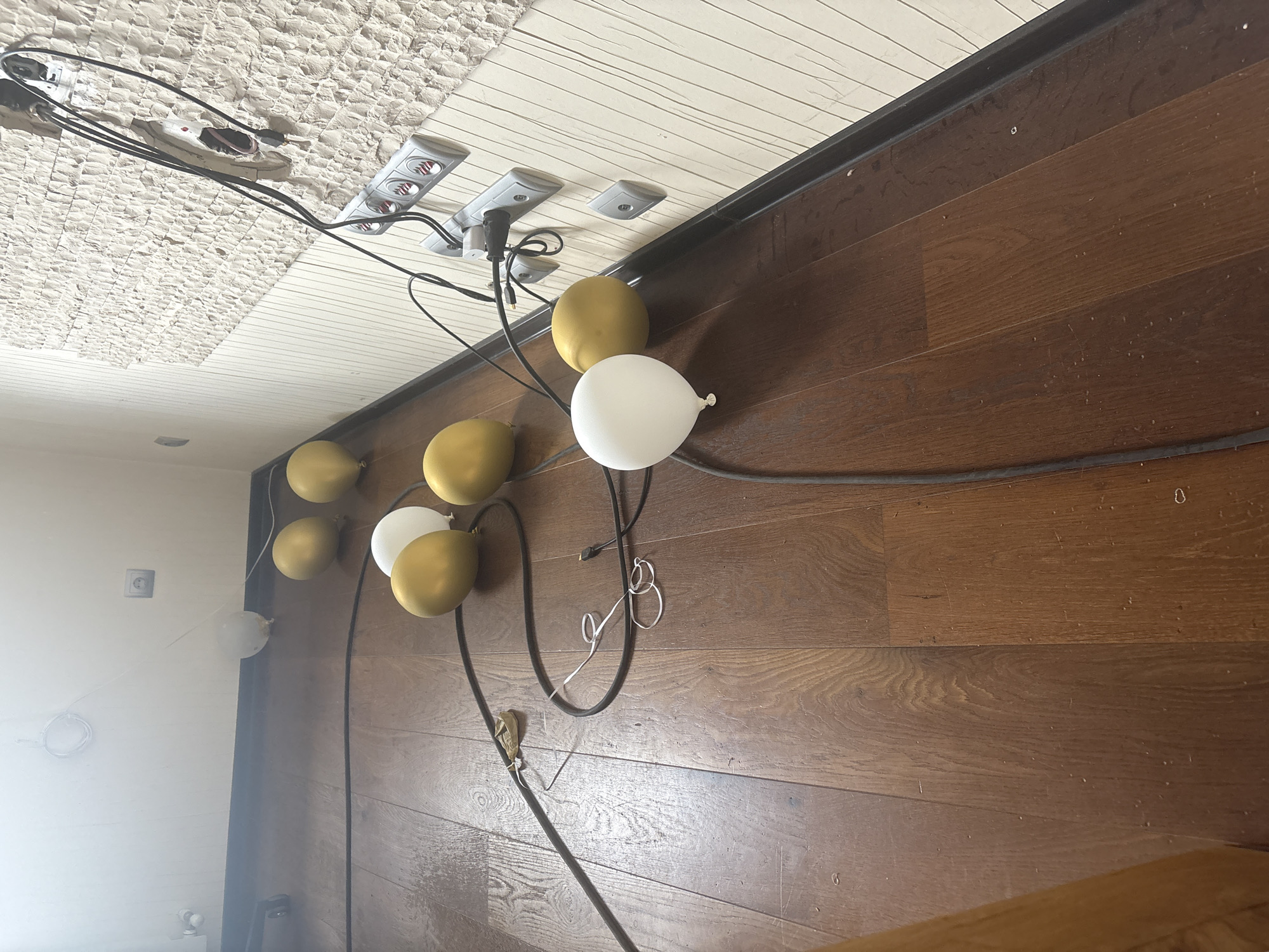 The image shows a dimly lit room with a dark hardwood floor. Several balloons, a mix of gold and white, are scattered across the floor, some of them deflated or partially deflated. Black electrical cords are strewn among the balloons and stretch towards a wall covered in a textured beige wallpaper. The wall also features a series of electrical outlets and what appears to be a power strip. In the background, there is a hint of a white wall with a light switch and a circular object mounted on it. The overall impression is one of a room that has recently been used for a celebration or event, with remnants left behind. There are no people visible in the image. The location is in the city of Orekhovka, Russia.