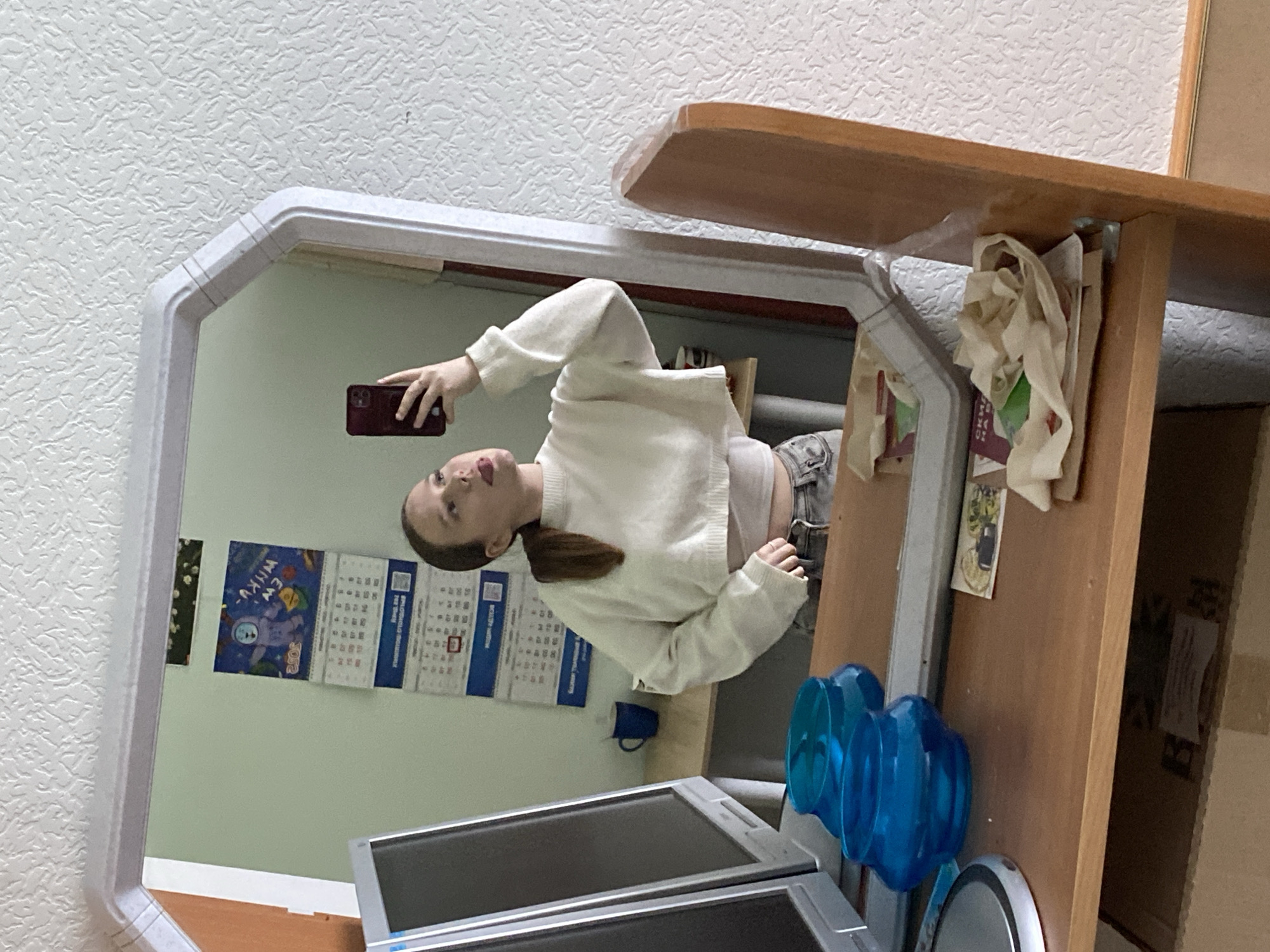 A woman with dark hair is visible in a mirror, holding a smartphone and taking a selfie. She is wearing a white sweater and appears to be in a room with light green walls. Calendars are visible behind her in the mirror. The room contains a computer monitor, blue water cooler jugs, and a wooden desk or shelf. The location of the photograph is likely in Chaikovsky, Russia, due to elements on the calendar.