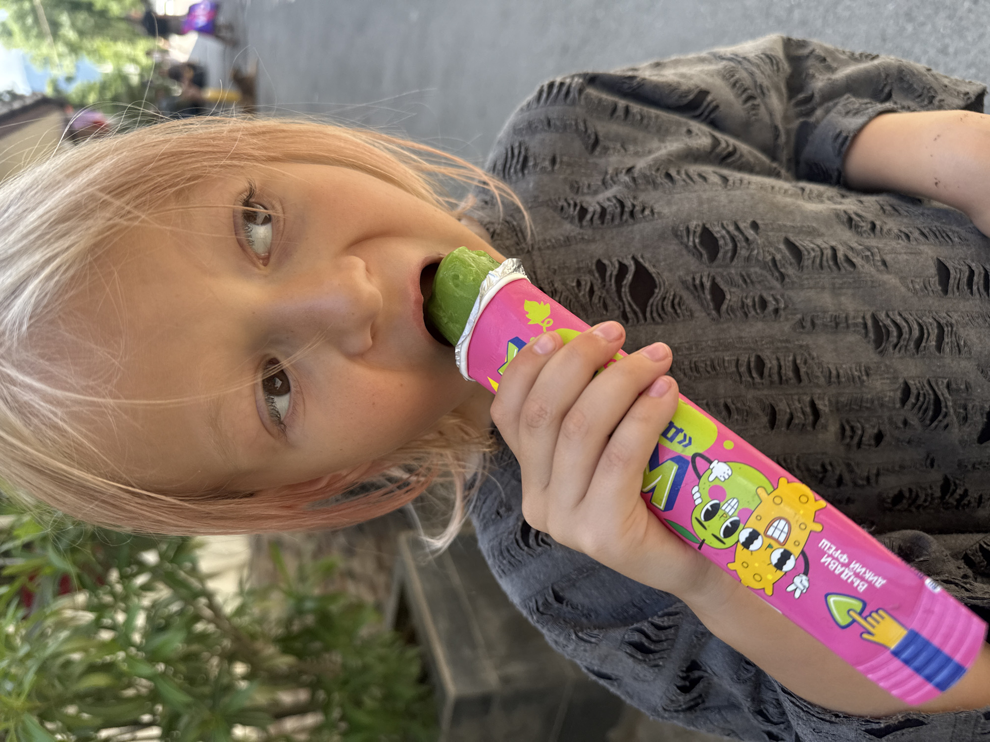 In this close-up, vertically oriented image, a young child with light blonde hair is leaning in to take a bite of a green popsicle. The child's mouth is open, and their eyes are wide, looking towards the camera. The popsicle is encased in a bright pink wrapper decorated with cartoon characters and a cartoon shovel. The child is wearing a dark gray, distressed t-shirt with ripped detailing. The background is blurred, but appears to be an outdoor setting with greenery and some structures visible. The lighting suggests it is daytime, and the overall mood is one of casual enjoyment. Visible text on the popsicle wrapper is in a non-English alphabet.