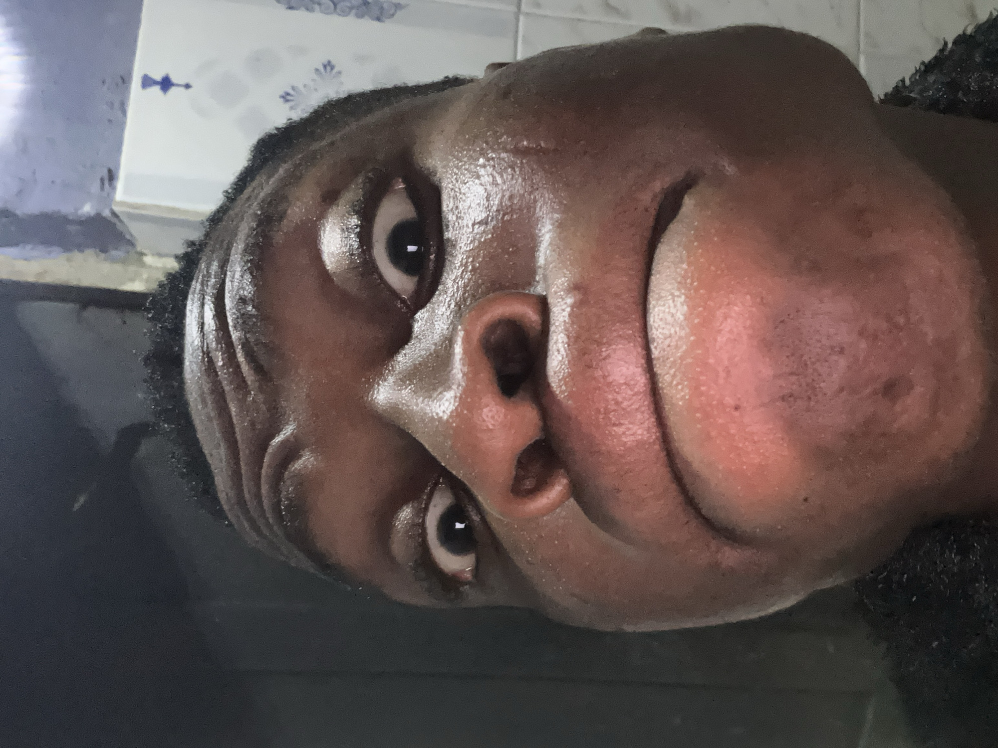 The image shows a close-up, distorted view of a dark-skinned person's face. The person is looking directly at the camera. The face appears shiny, possibly due to sweat or oil. The background is partially visible, showing what appears to be a wall with some decorative tile designs. The location is specified as Unknown, Unknown.