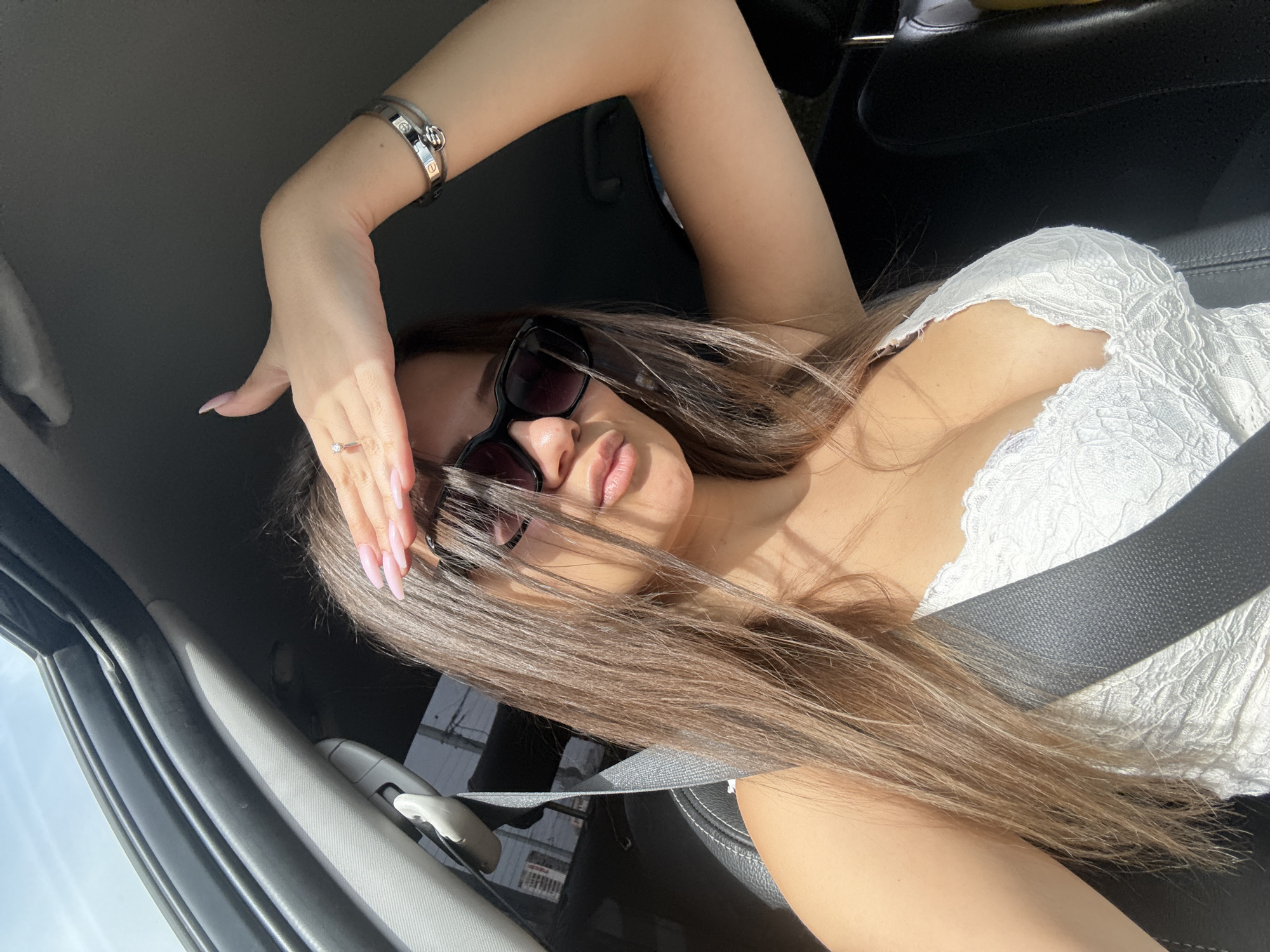 A woman is taking a selfie inside a car. She is wearing a white lace top, sunglasses, and a bracelet. Her hair is long and brown. She is holding her hand up to shield her eyes from the sun. The car is parked. The exact location in Sofia, Bulgaria is not identifiable from this image.