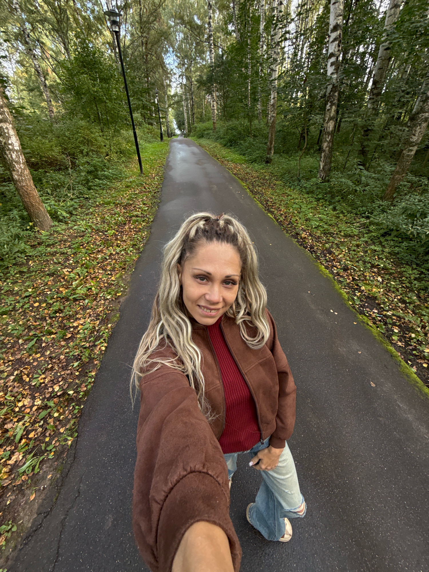 In Rusanovo, Russia, a woman with blonde dreadlocks takes a selfie on a paved path through a forest. She is wearing a brown jacket over a red ribbed shirt and light-wash ripped jeans. The path is lined with trees and green foliage, with fallen leaves scattered on the ground. A lamppost is visible to the left of the path. The scene is outdoors, suggesting a park or wooded area.