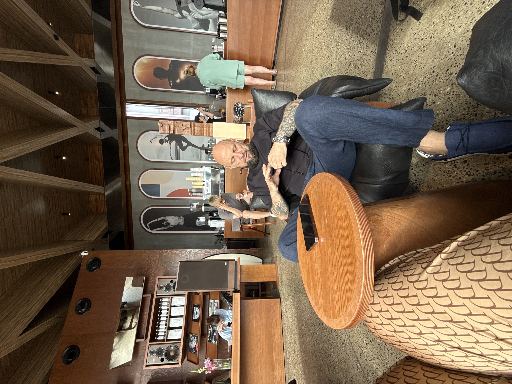 In an indoor cafe, a bald man with a beard and tattoos on his arms sits in a black leather armchair, looking down at his phone. He wears a black t-shirt and blue pants. In front of him, a small round wooden table holds a black smartphone. The cafe features a rustic wooden ceiling with exposed beams and decorative panels. The walls are adorned with framed artwork depicting dancers and abstract shapes. A wooden counter with coffee machines and products is visible in the background, where a woman in a green outfit stands, and another person operates the coffee machine. To the left, a woman in a blue shirt leans over a wooden counter, possibly working. The floor is a speckled grey concrete.