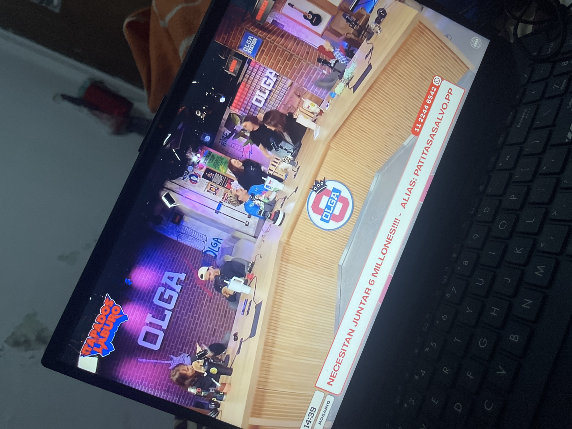 The image displays a laptop screen showing a live stream of a radio or television show. The program appears to be called "OLGA" and features a group of people seated around a wooden desk. The background is decorated with brick patterns and various posters or graphics, including guitars. A prominent banner at the bottom of the screen reads "NECESITAN JUNTAR 6 MILLONES!!! - ALIAS: PATITASASALVO.PP" with a phone number "11 2244 6542" and a WhatsApp icon. There is also a timestamp "14:39" and the text "ROSARIO" visible. The overall setting is that of a studio. The laptop is placed on a dark keyboard, suggesting the viewer is at a desk or computer. There is no visible indication of the location being Santa Ana, Venezuela.