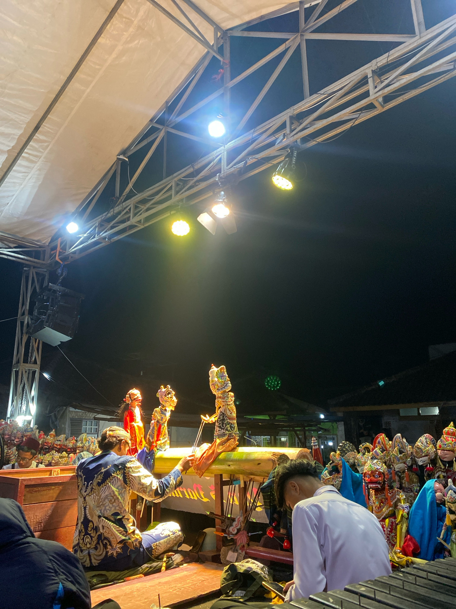 This is a medium shot image capturing a traditional Wayang Kulit (shadow puppet) performance in the city of Sugihmukti, Indonesia. The scene takes place outdoors at night, illuminated by stage lighting. In the foreground, a puppeteer dressed in a blue and gold ornate garment is seated on the floor, actively manipulating a puppet with his right hand. He is facing away from the camera, towards a flat wooden surface where the puppets are being presented.
To the left of the main puppeteer, another figure in similar traditional attire is also engaged with puppets. In the background, a collection of elaborate Wayang Kulit puppets are displayed, showcasing their detailed craftsmanship and vibrant colors. The stage is set up with a wooden platform and a translucent screen, typical for shadow puppet performances. A metal truss structure with multiple spotlights is visible above the stage, providing the necessary illumination for the show. The overall environment suggests a festive or cultural event, with the dark night sky serving as a backdrop.
