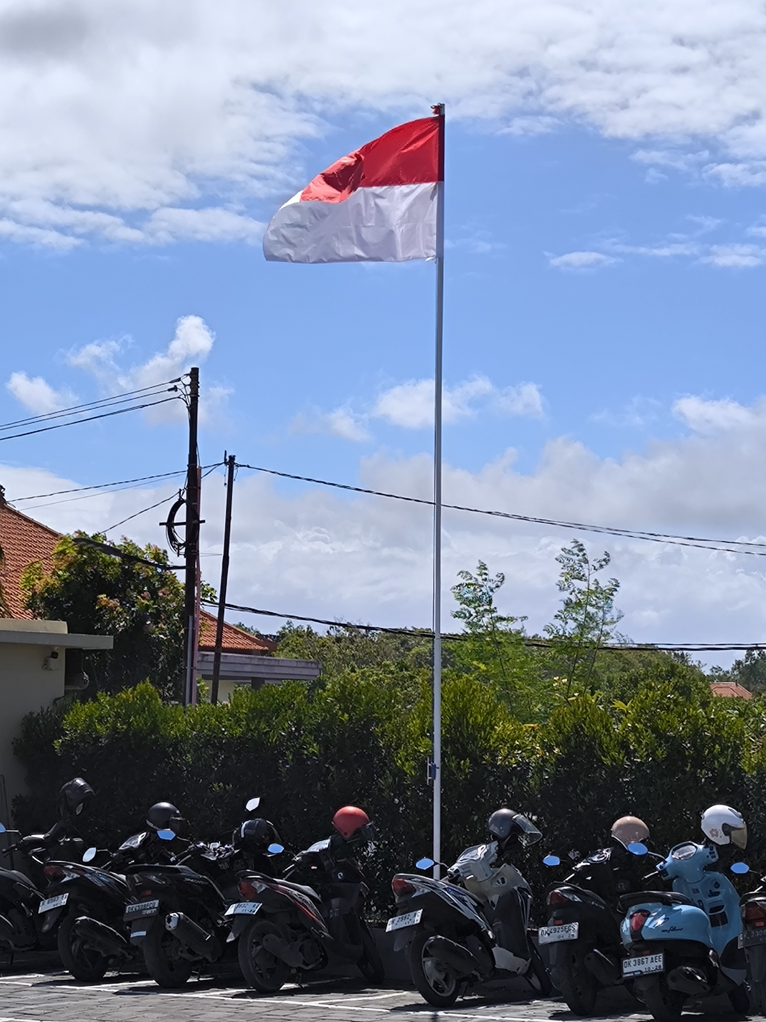 The image shows a row of parked motorbikes in front of a building with a tiled roof and shrubbery. Above the building and shrubbery, a flag with a red stripe over a white stripe is flying on a pole against a blue sky with white clouds. Power lines are visible in the background. The city location is Unknown.