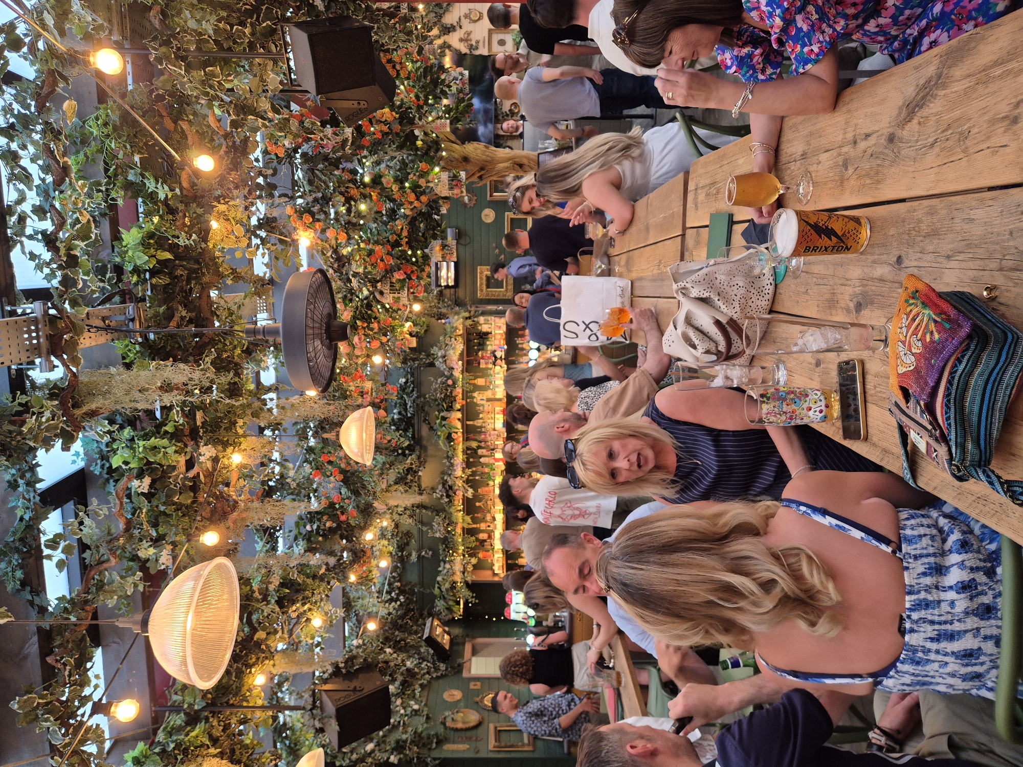 The image depicts an indoor scene, likely a restaurant or bar, in Cardiff, United Kingdom. Several people are seated at a long wooden table, drinking and socializing. The table is crowded with drinks, glasses, and personal belongings such as purses and phones. Some individuals are in close conversation, while others appear more relaxed. The overhead space is heavily decorated with artificial foliage, including leaves, flowers, and strings of lights. A fan is also visible suspended from the foliage. The lighting is dim, creating a warm and festive ambiance. The background features a bar area stocked with bottles and a wall adorned with decorative items and framed pictures.