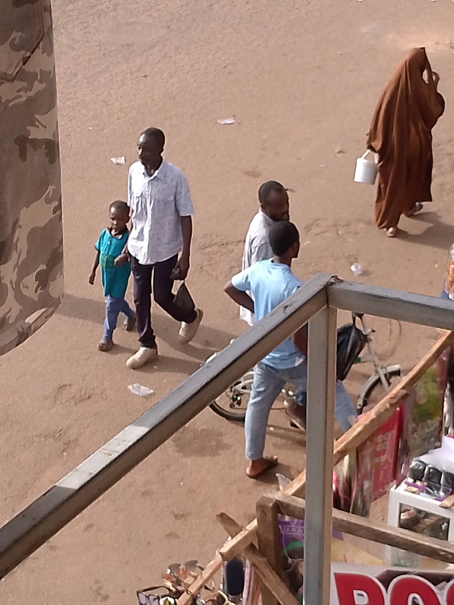 A group of people are walking outdoors on a dirt path. A man wearing a light-colored, short-sleeved collared shirt and dark pants walks with a young child dressed in a turquoise t-shirt and light blue pants. Behind them, two other men are walking, one in a white shirt and the other in a light blue t-shirt. Further away, a person in a long brown garment, likely a woman wearing a hijab, carries a white bucket. A bicycle with a black bag attached to the handlebars is stationary nearby. The ground is a light brown dirt surface with scattered debris. A portion of a wooden structure with a metal railing is in the foreground, suggesting a raised walkway or balcony overlooking the scene. The overall atmosphere appears to be a marketplace or a busy pedestrian area.