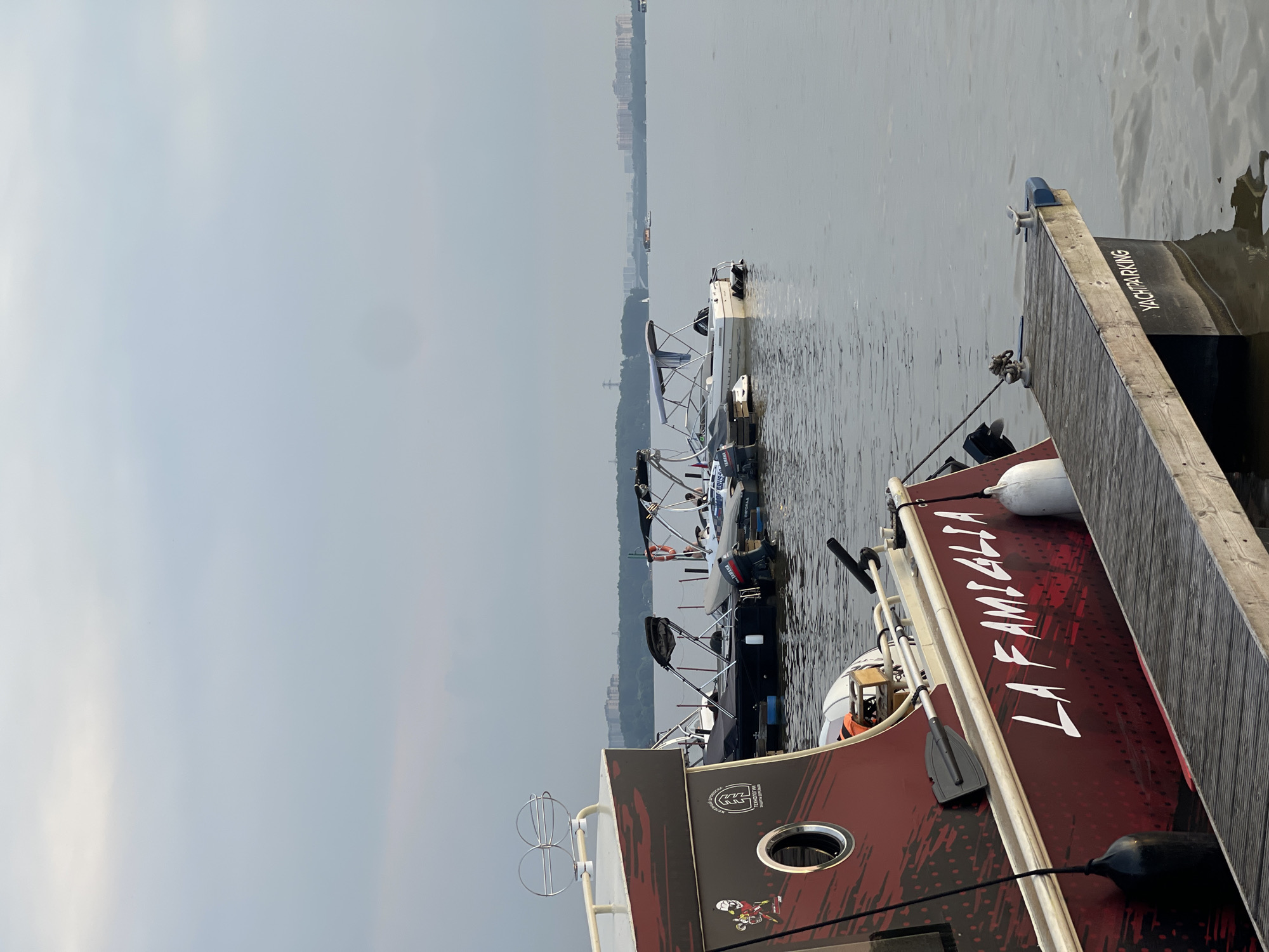 This is a photograph taken from a dock at the city of Novoseltsevo, Russia. The image shows several boats moored at the dock, with a wide expanse of water stretching out to the horizon. In the distance, the cityscape of Novoseltsevo is visible, characterized by buildings and trees. The sky is overcast, with a soft, diffused light.
In the foreground, a large, dark red boat is prominently displayed. It has a graphic design on its hull with the words "LA FAMIGLIA" in white lettering. A wooden dock extends from the right side of the image towards the boat. The dock has a sign on its side that reads "YACHTPARKING". There are fenders hanging from the side of the large red boat.
Behind the foreground boat, several smaller boats are docked. These appear to be speedboats and fishing boats, some with outboard motors. One of these boats has a Yamaha engine. There are also lifebuoys visible attached to the boats. The water is calm, with gentle ripples. The overall atmosphere of the image is peaceful and serene.