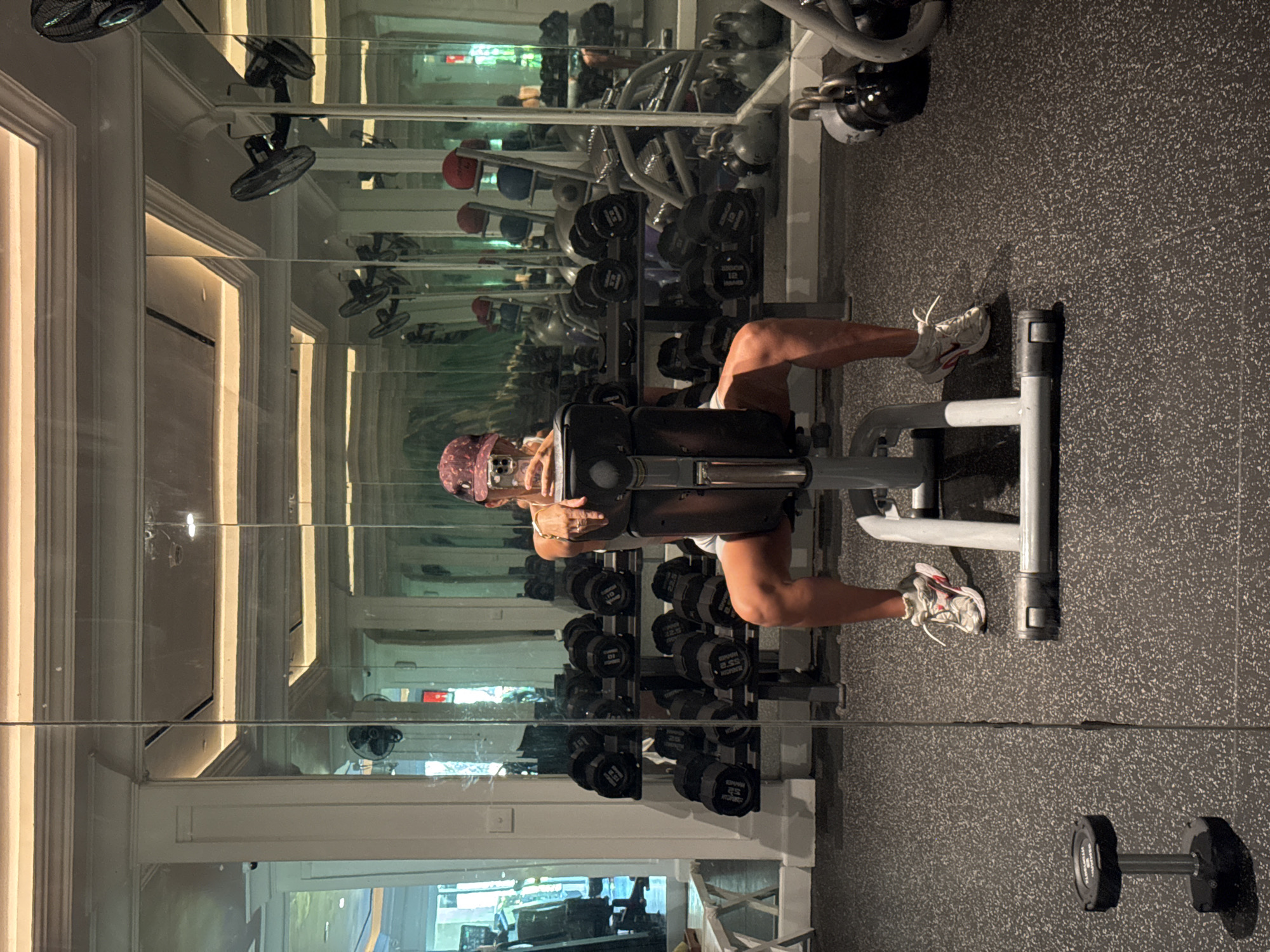 A person is sitting on a leg extension machine in a gym. The gym is located in Kuta Selatan, Indonesia. In the background, there are rows of dumbbells and other gym equipment. The person is wearing athletic shoes and a hat. The image is taken from a mirrored wall.