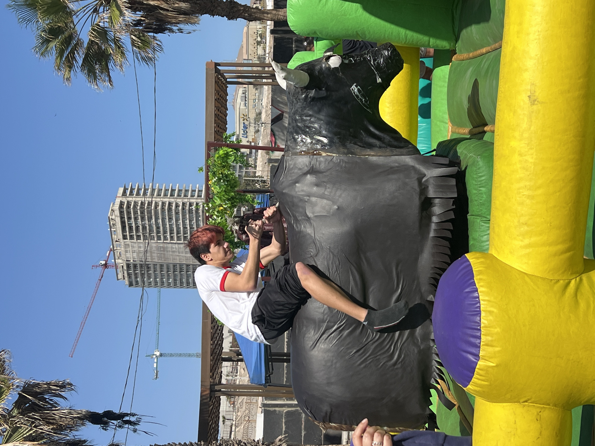 A young man with reddish hair is sitting on a mechanical bull, part of an inflatable rodeo ride, in an outdoor setting in Arica, Chile. He is wearing a white t-shirt with red trim and black shorts. The mechanical bull is predominantly black with white horns. The inflatable surrounding the bull has green, yellow, and purple sections. The background shows a construction site with cranes, a tall building under construction, and palm trees against a clear blue sky. A partial view of a building with the "Lider" sign is visible.