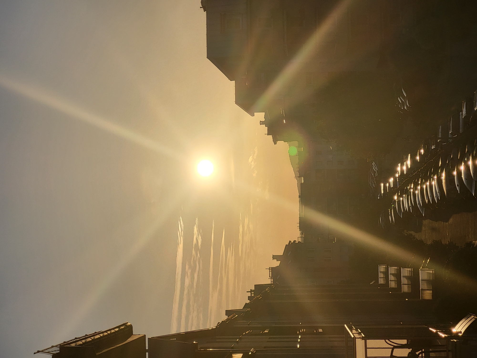 The image is a vertical, medium shot of an urban landscape during sunset. The sun, a bright white orb, is positioned in the upper center of the frame, emitting strong sun flares that extend across the sky and through the buildings. The sky is a gradient of pale orange and yellow. Several buildings, appearing to be apartment complexes, line both sides of a street or courtyard. The buildings on the left are silhouetted against the sky, with balconies visible. The buildings on the right are also silhouetted and appear to be of similar height. In the foreground and extending into the middle ground, a row of parked cars is visible, their surfaces reflecting the bright light from the sun. The overall impression is a residential area bathed in the warm, dramatic light of a setting sun. The exact location is Unknown, Unknown. There are no people visible in the image.