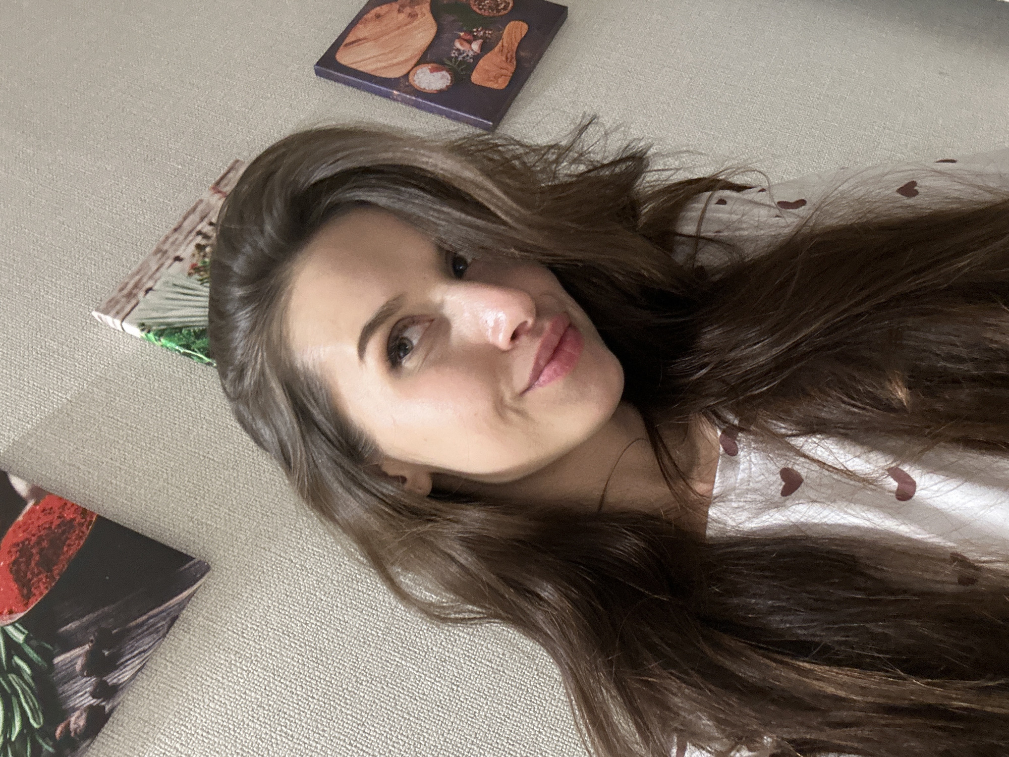 A woman with long brown hair is lying on a light-colored, textured surface, possibly a sofa or bed. She is wearing a white top with small red heart patterns. Three square objects with images on them are positioned around her head. One has a cooking-related theme with bread or ingredients, another has a red food item with rosemary, and a third has a picture with green and white elements. The woman is looking directly at the camera with a slight smile.
The location is not identifiable as being in Yekaterinburg, Russia, based on the provided image.