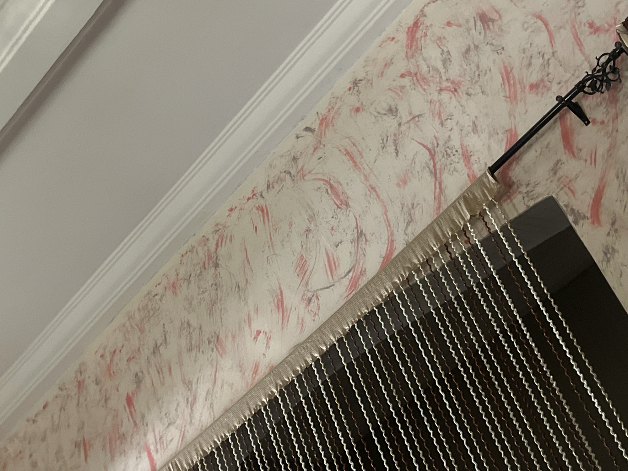 The image shows a close-up of a wall with textured wallpaper featuring a pattern of red and gray brushstrokes on a cream background. A white crown molding is visible along the top edge, separating the wall from a plain white ceiling. In the foreground, a beaded curtain with thin, wavy strands in brown and white hangs across the lower right portion of the frame. A black curtain rod with an ornate decorative finial is mounted on the wall above the curtain. The scene appears to be indoors, likely in a residential setting. There are no people or animals present in the image. The lighting suggests it might be taken during the day with artificial light, or in the evening. The specific location in Jalingo, Nigeria cannot be determined from the visual information provided in the image.