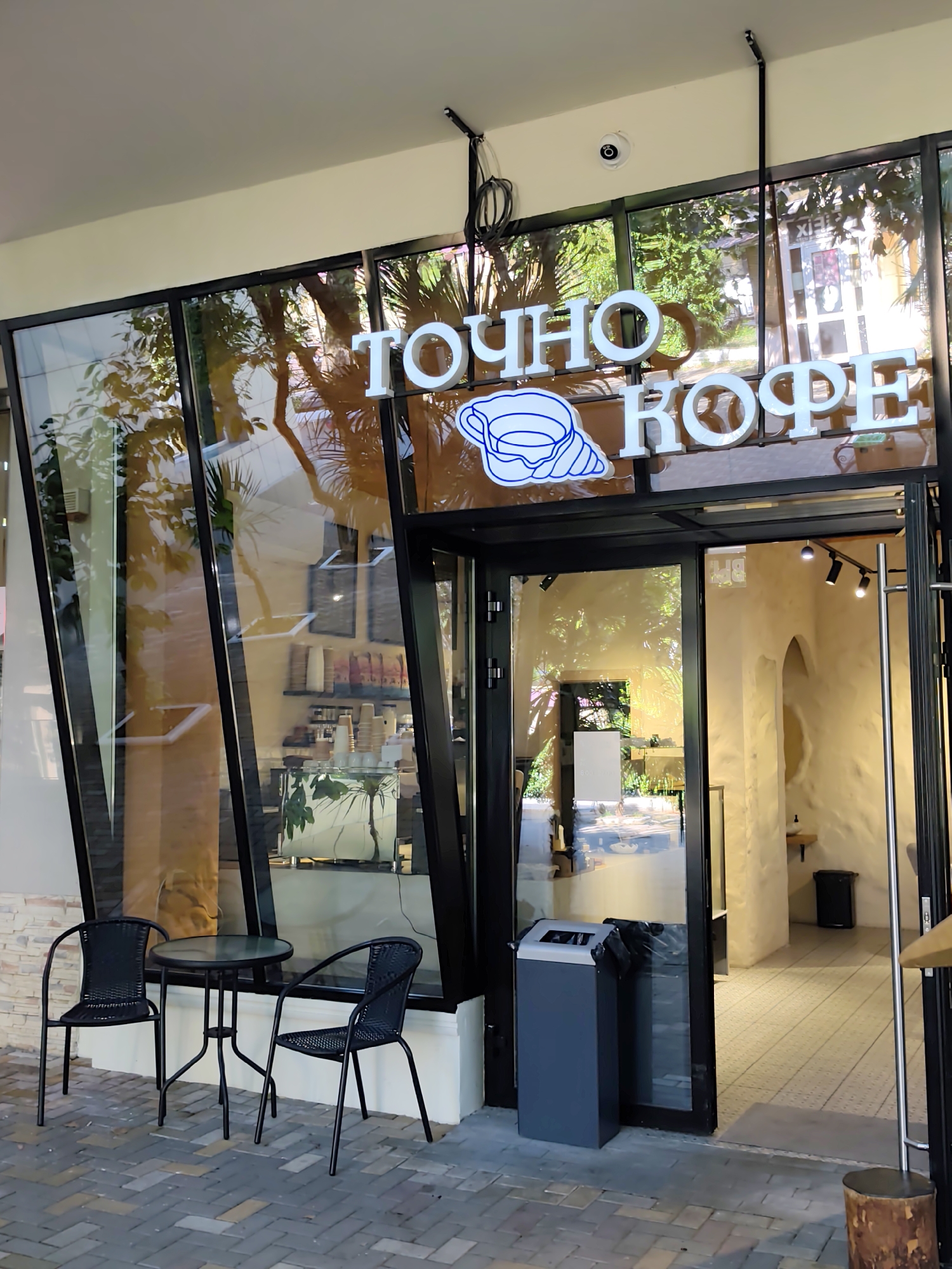 The image shows the exterior of a coffee shop named "ТОЧНО КОФЕ" (TOCHNO KOFE) located in Sochi, Russia. The shop has a large glass facade with a neon sign displaying the name. In front of the shop, there is a small outdoor seating area with a round table and two black chairs. To the right of the entrance, there is a gray trash bin. The entrance is an open glass door leading into the interior of the coffee shop, which features white walls and arched doorways. The flooring outside the shop is paved with interlocking bricks, and there are some potted plants visible. The overall scene depicts a modern and inviting coffee shop.