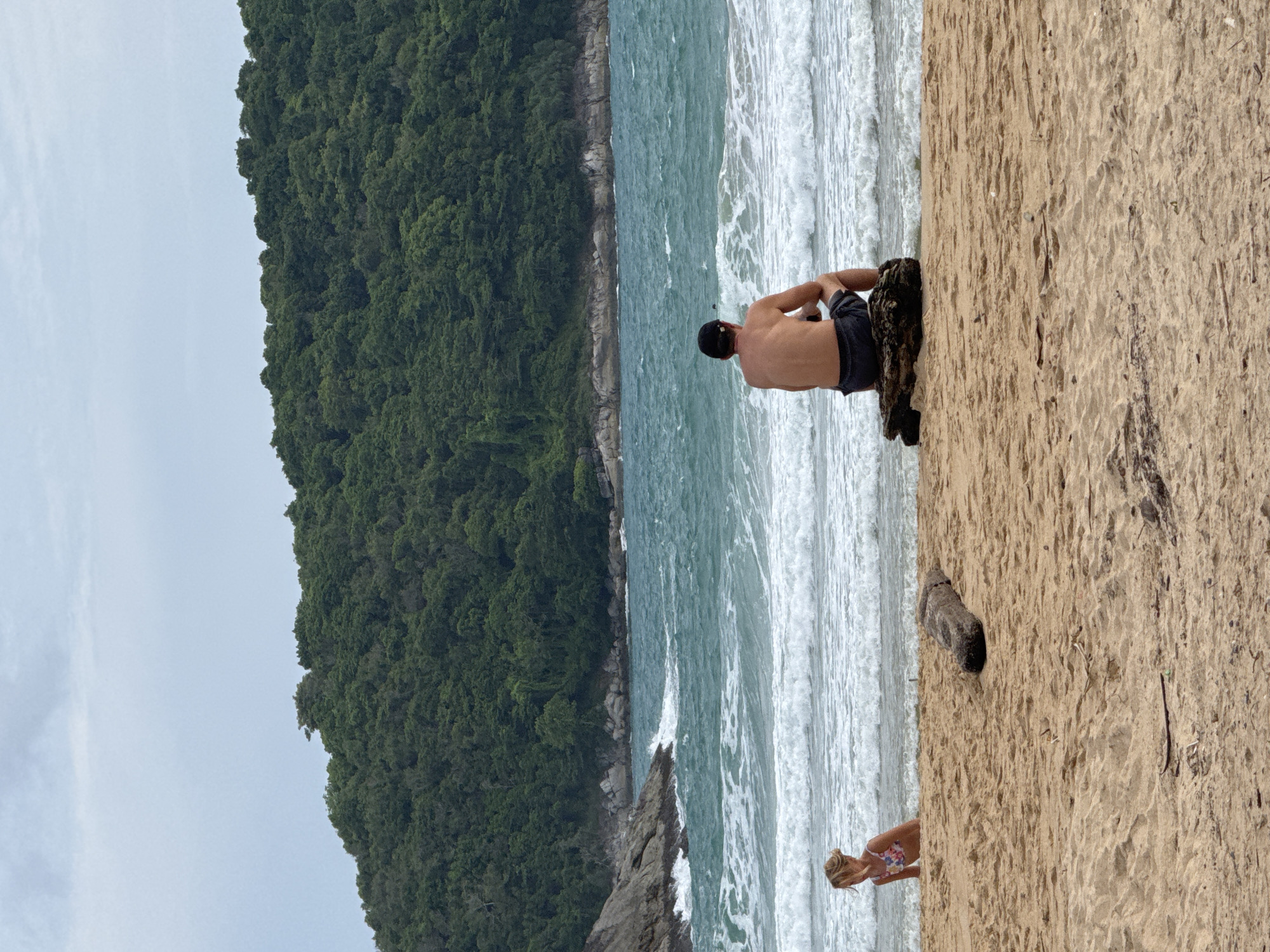 The image depicts a scenic beach scene in Rawai, Thailand. The primary subjects are two people on the sandy shore. In the foreground, a person in a floral swimsuit is partially visible, looking towards the ocean. Further back, a shirtless man wearing dark shorts and a baseball cap sits on a large dark rock, facing the sea. His posture suggests he is relaxed or contemplative.
The setting is a tropical beach with a dense, green forest covering a hillside that rises steeply from the rocky coastline. The ocean is a vibrant turquoise, with white waves crashing onto the shore, indicating a moderate surf. The sand is light brown and appears dry in most areas. A few pieces of driftwood are scattered on the beach. The sky is overcast and a pale grey, suggesting it is either early morning or late afternoon, or the weather is cloudy. The overall mood is tranquil and natural. There is no visible text in the image.