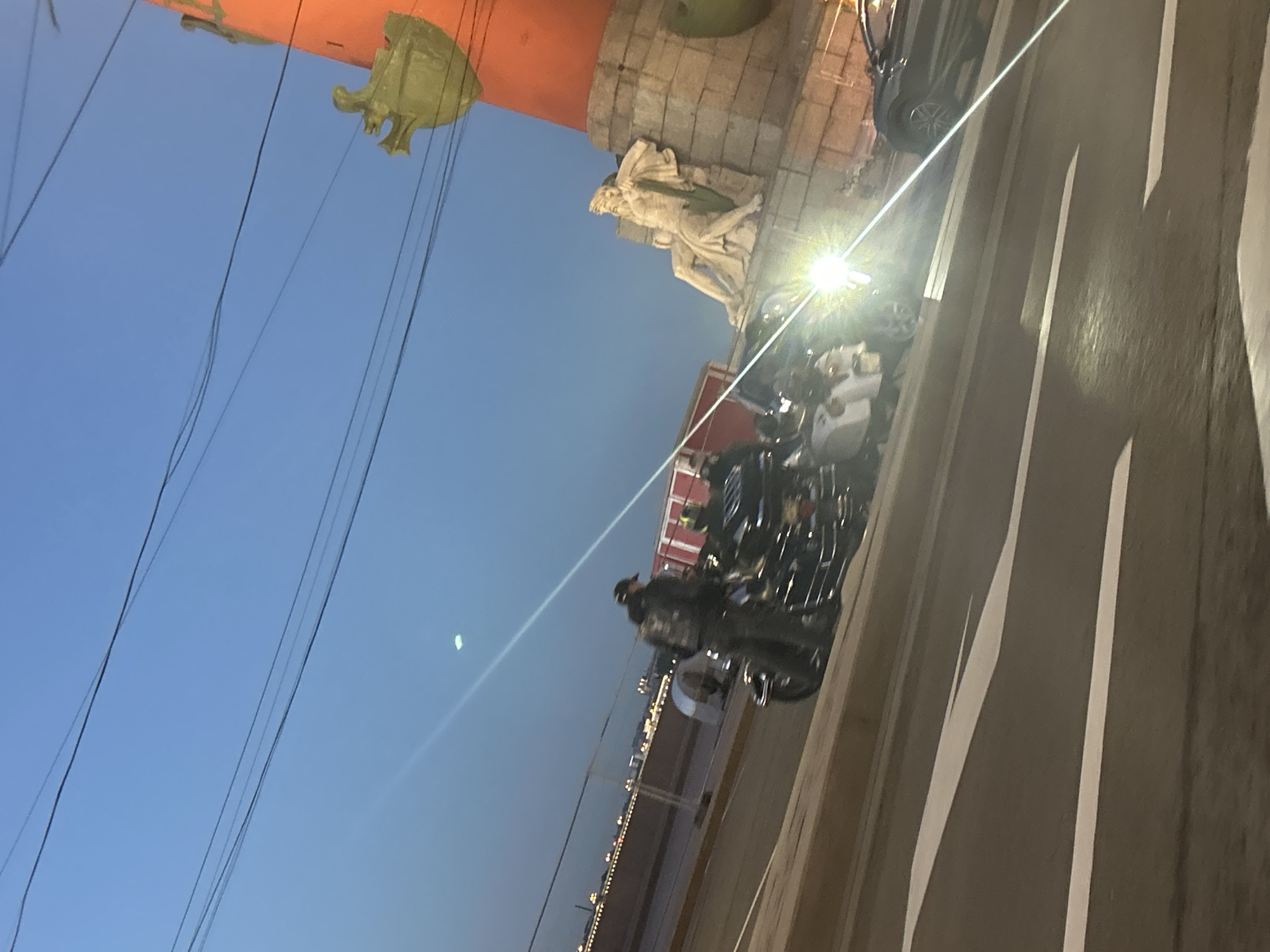 The image depicts a group of motorcycles parked on a street next to a large, ornate column. The column is orange at the top and features a golden sculpture, possibly a ship's prow, and a classical statue. The sky is a clear, deep blue, suggesting it is either early morning or late evening. A bright light, likely from a streetlight or a nearby building, illuminates the scene, creating strong highlights and shadows.
The motorcycles are the primary focus in the lower part of the image, with several of them appearing to be custom or cruiser style. One rider is visible on a motorcycle, wearing a helmet and dark clothing, suggesting they are either about to leave or have just arrived. The street appears to be paved with asphalt and has white lane markings.
In the background, across the street, there are buildings with illuminated windows and lights stretching along the horizon, further indicating a nighttime or twilight setting. The overall atmosphere is somewhat dramatic due to the lighting and the imposing presence of the column. There is no visible text in the image.