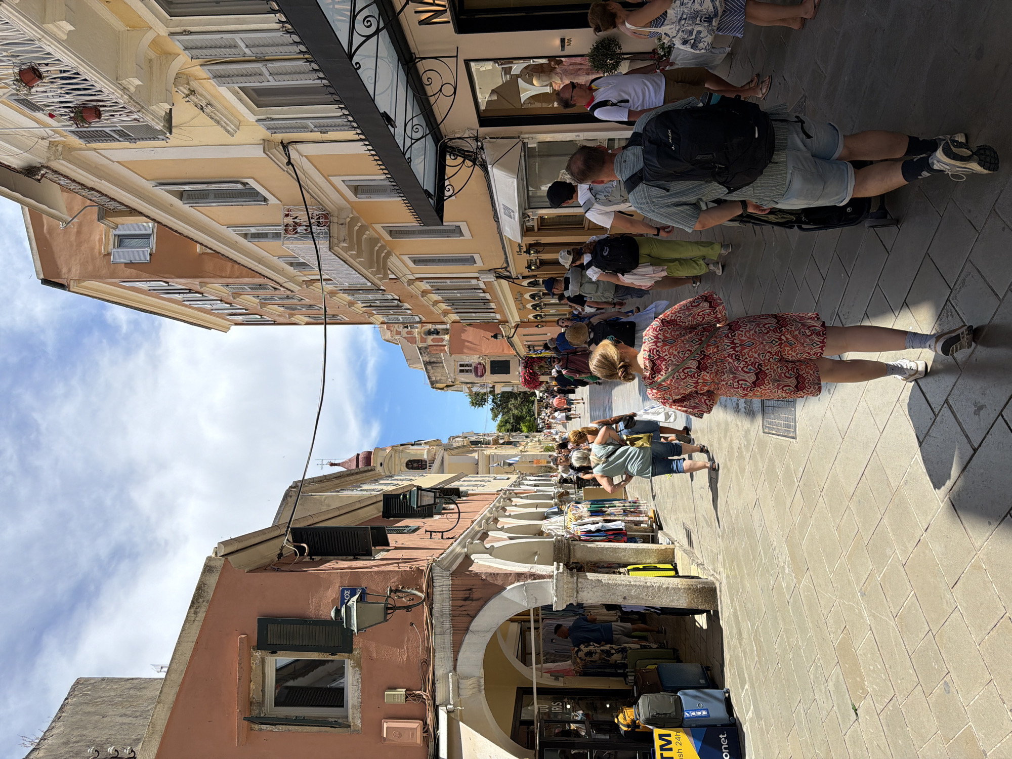 This media file, viewed after being rotated counter-clockwise, captures a lively pedestrian street scene in Corfu, Greece, during a bright daytime with a clear blue sky and scattered white clouds. The narrow street is paved with light-colored square tiles and is bustling with people.
Multi-story buildings line both sides of the street. On the left, the buildings are painted in shades of light yellow and a warm orange-brown, featuring traditional shuttered windows and some balconies with hanging flower pots. An arched passageway at ground level leads to a shop, above which "IRIS" is visible, along with a blue street sign displaying "ΣΟΣ". Further down the street on this side, a church spire or bell tower can be seen.
On the right side, buildings are primarily light yellow and cream, housing various shops at ground level, some with dark awnings and window displays, including mannequins. Numerous people are walking in both directions, appearing to be a mix of locals and tourists, dressed in casual summer clothing such as shorts, dresses, and t-shirts. In the foreground, a person in a red and white patterned dress walks away, while next to them, another individual in denim shorts, a striped shirt, and a black backpack (with "VAUDE" visible) pushes a trolley. An ATM sign with "TM" and "English 24h" text is visible near a shop on the right. The scene depicts a vibrant and active commercial area within a historic European town.