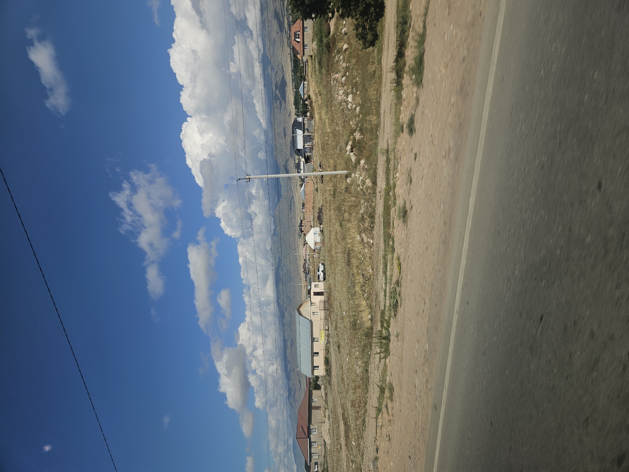 In Ornek, Kyrgyzstan, a suburban landscape unfolds under a bright blue sky dotted with fluffy white clouds. A paved road, with a white line marking its edge, runs across the foreground, suggesting it's a roadside view. Beyond the road, a dry, grassy field stretches out, interspersed with scattered rocks. Several houses with different architectural styles are visible. On the left, a house with a red-tiled roof and beige walls stands behind a low fence. To its right, a lighter-colored house with a blue-grey metal roof features a sign in a foreign language. Further back, more houses and structures, including a yurt, are scattered across the terrain. A single utility pole with wires stretches across the scene. In the distance, a range of mountains forms a hazy backdrop, their slopes appearing dry and arid. The overall impression is one of a rural or semi-rural community with a mix of traditional and modern housing, set against a vast natural landscape.