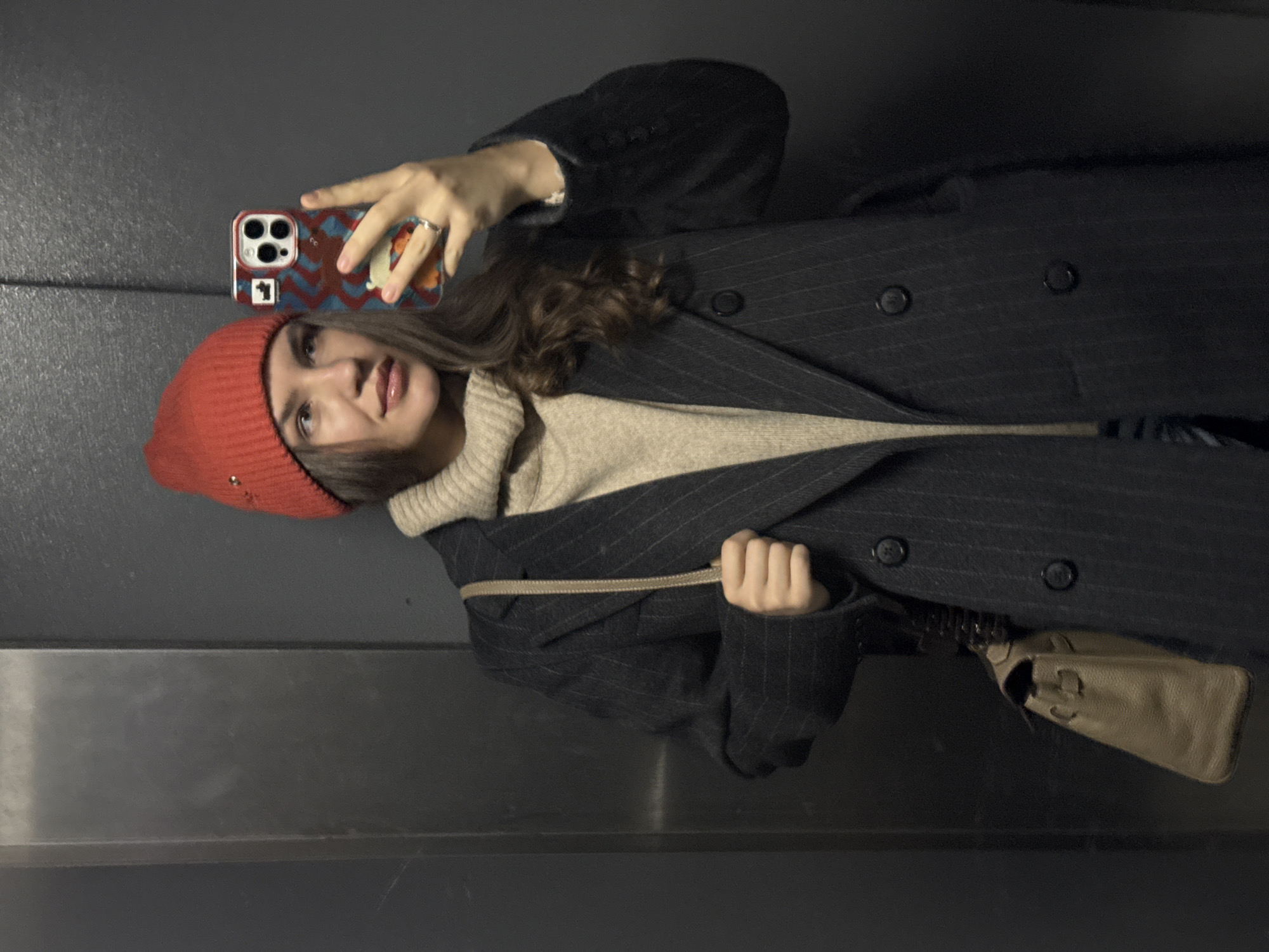 A young woman is captured in a selfie, holding a smartphone with her right hand. The image is presented rotated ninety degrees to the left, showing the woman as if lying on her back, but she is likely standing upright, taking a mirror selfie or direct selfie against a dark, featureless wall.
The woman has medium-length, dark brown wavy hair, and is wearing a red knit beanie or beret. She has a subtle smile on her face, with soft makeup including what appears to be glossy lips. Her attire consists of a light beige or off-white ribbed turtleneck sweater, layered under a dark, possibly navy or black, pinstripe overcoat. The overcoat features large dark buttons.
She holds a smartphone with a distinctive, colorful patterned case, featuring a red and blue zigzag design and what looks like an orange-brown animal character on the right side. A silver ring is visible on her right ring finger. A light brown or tan leather handbag with a strap is visible, slung over her arm or shoulder.
The background is a dark, plain wall, possibly metal or painted, with a subtle horizontal seam or panel line. The lighting appears to be artificial and somewhat subdued, common for an indoor setting like a hallway or an elevator, consistent with the provided context of Moscow, Russia. There is no visible text in the image. The overall mood is casual and contemporary.
