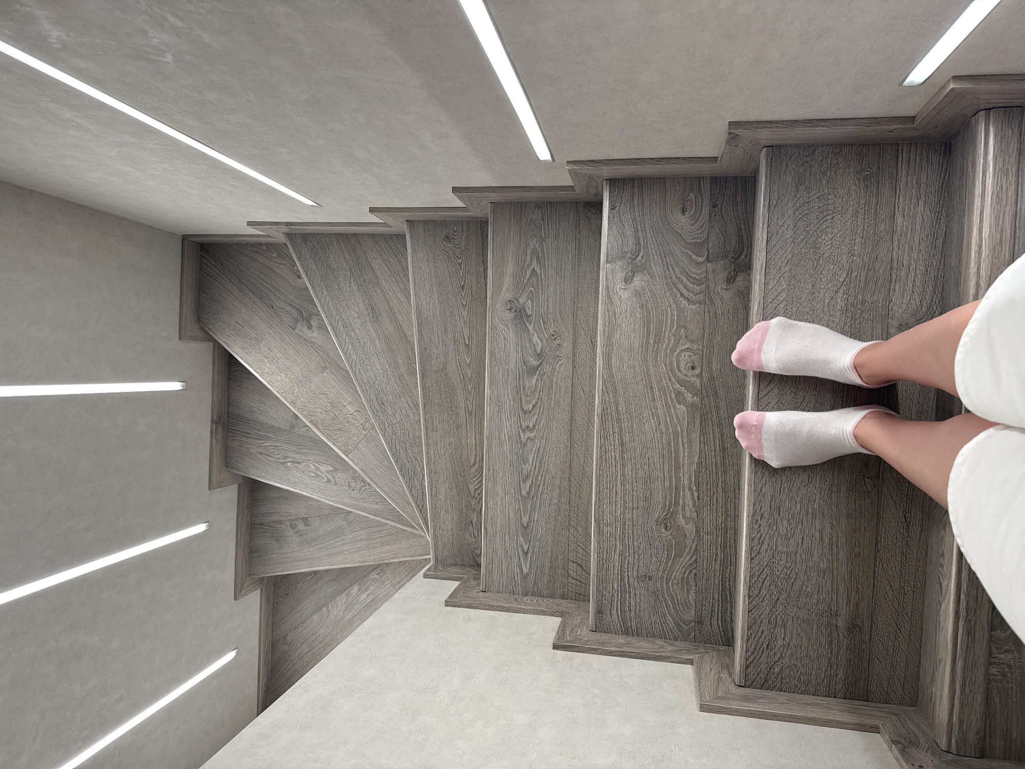 The image provides an overhead perspective looking down a modern, winding staircase within an interior setting. A person's bare legs and feet, wearing white socks with pink accents on the toes and heels, are positioned on one of the top steps on the right side of the frame. A hint of a white garment, possibly a robe or towel, is visible near the person's right leg.
The staircase is constructed from dark grey wood, showcasing a distinct grain pattern. The steps are thick and appear to be individual, possibly cantilevered, forming a smooth, curved descent. The design gives the impression of a floating or sculptural element.
The surrounding walls and ceiling are a uniform light grey, featuring a subtle texture that suggests concrete or a similar finish. Strips of bright, linear LED lighting are integrated into both the ceiling and the walls, illuminating the staircase and contributing to a sleek, contemporary atmosphere. These light strips create sharp lines that contrast with the muted grey tones and the organic patterns of the wood. The floor at the base of the stairs is also a light grey, textured surface, seamlessly blending with the walls. The overall aesthetic is minimalist and modern, emphasizing clean lines and functional lighting. There is no visible text in the image.
