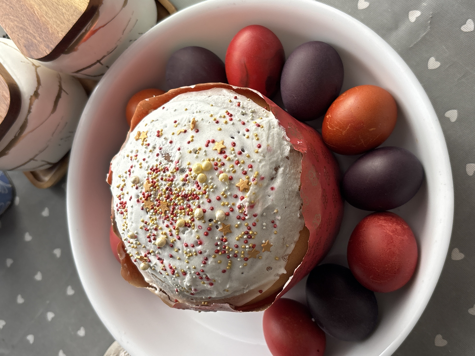 A high-angle shot shows an Easter cake and dyed eggs on a white plate. The cake has a white frosting covered in colorful sprinkles and is sitting in a red paper liner. Around the cake are red, purple, and copper-colored eggs. In the background, partially visible, are white containers with wooden lids. The plate rests on a grey surface patterned with white hearts. The location is Фоки, Russia.