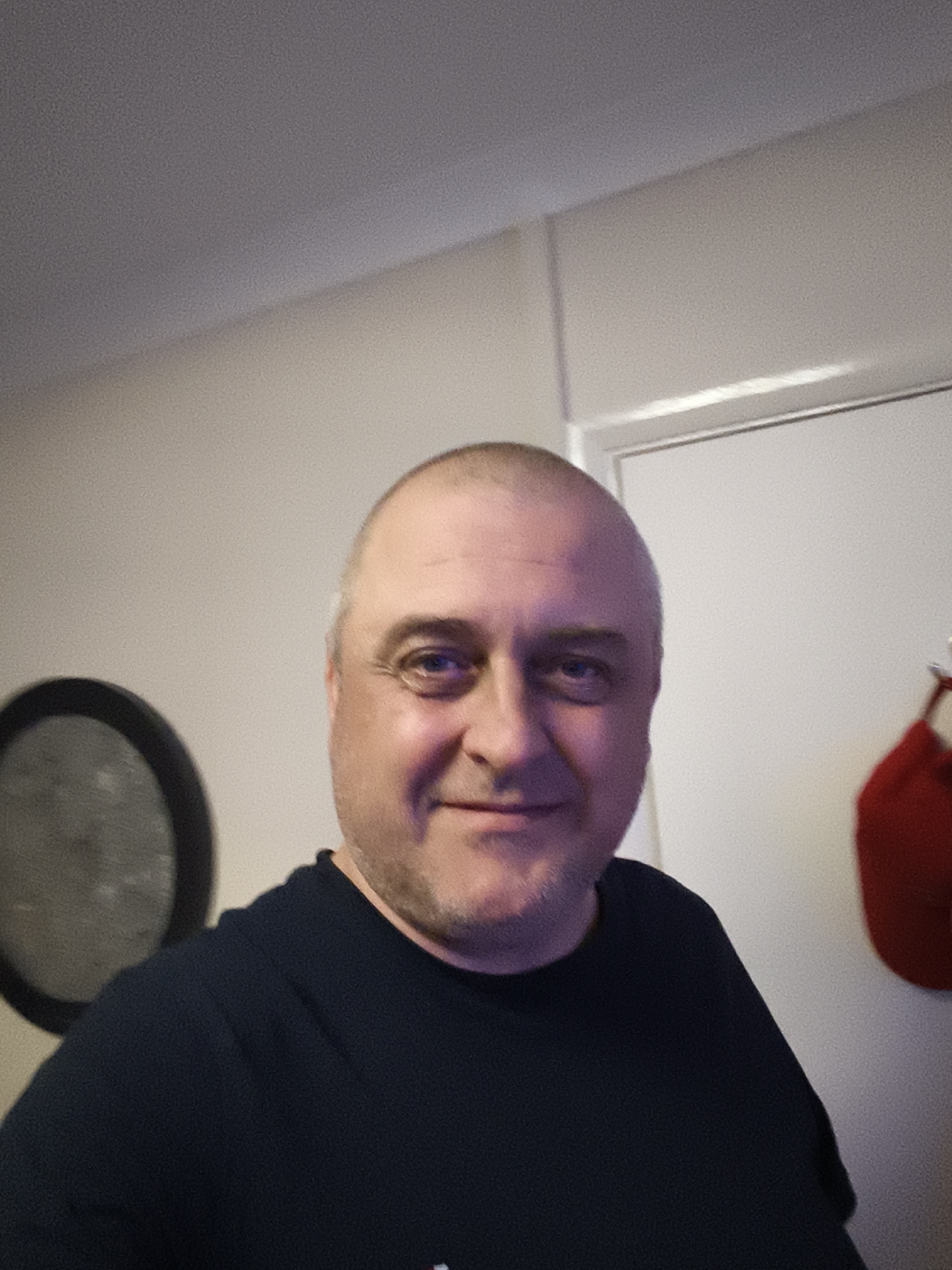 The image displays a close-up, slightly off-center portrait of a middle-aged man smiling indoors in Huntingdonshire, United Kingdom. He has light skin, closely cropped or shaved light-colored hair, and light stubble on his face. His eyes, which appear blue or grey, reflect a light source. He is wearing a dark, plain t-shirt, likely black or navy blue. His expression is friendly and content.
The background reveals light-colored walls, possibly beige or off-white, and a white door frame on the right side of the frame. To the man's left, a portion of a large, dark circular object with a thick rim is visible, possibly a dartboard or a clock. On the right, hanging on or near the door frame, is a bright red object, which might be a Santa hat or a small, decorative fabric item. The lighting is artificial, contributing to the reflections in his eyes. There is no visible text in the image. The scene suggests a casual, domestic environment.