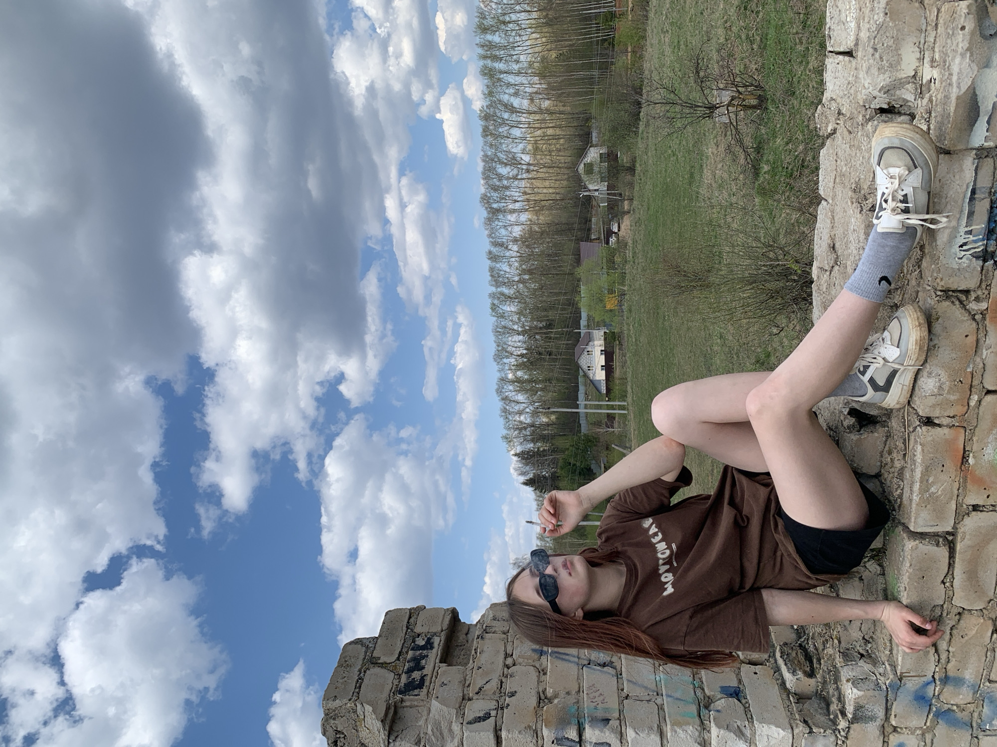 A young person lies on a brick structure in what appears to be an urban environment. They are wearing a brown t-shirt, black shorts, grey socks with the Nike logo, and white sneakers. The person is also wearing sunglasses and holding a lit cigarette in their right hand.
The brick structure shows signs of disrepair. The background consists of a landscape with trees and some buildings.
The location is Фоки, Russia. The sky is visible with clouds. The image has been rotated 90 degrees counterclockwise.